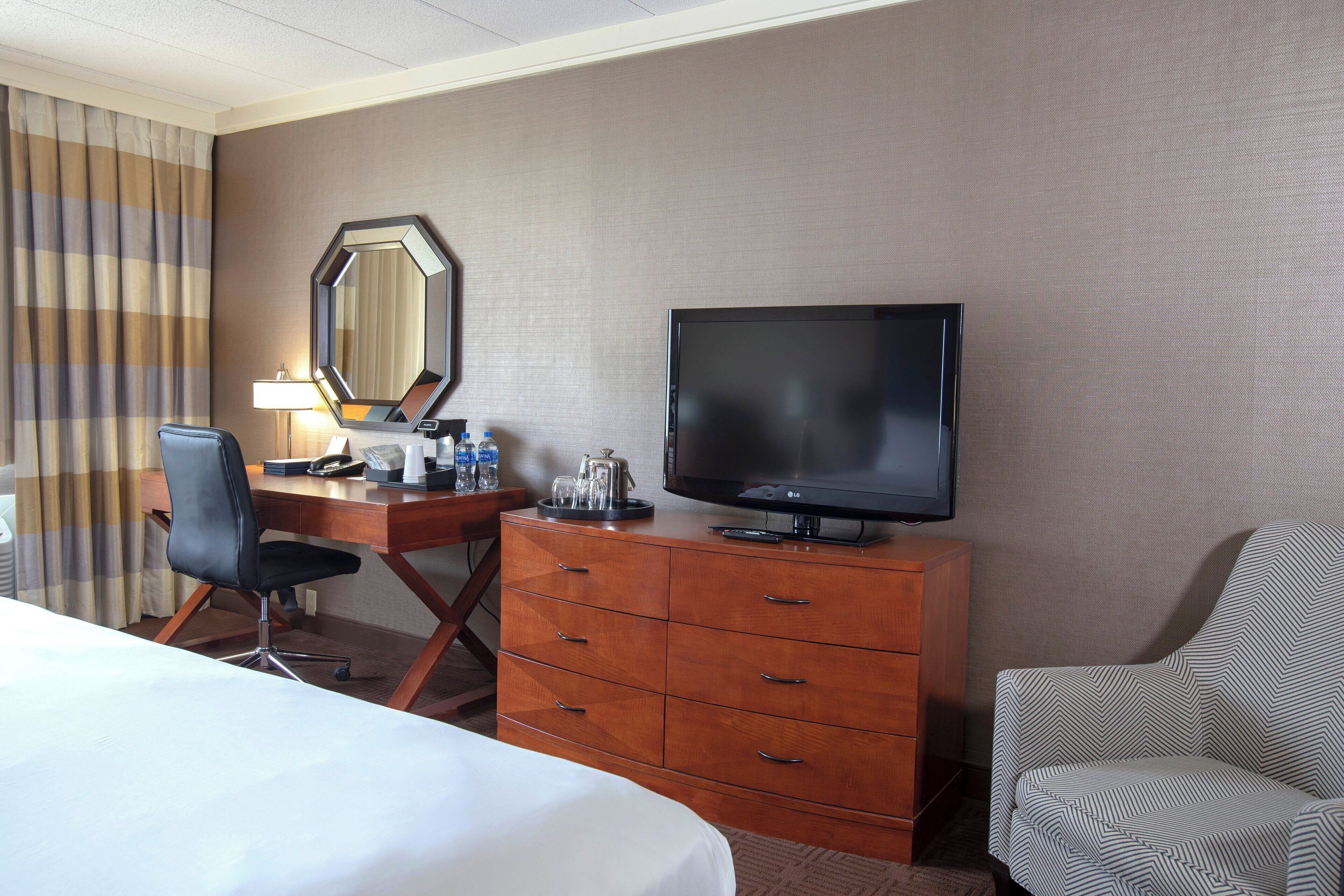 A hotel room with a bed, a desk with a chair, a television, a dresser, and a large mirror.