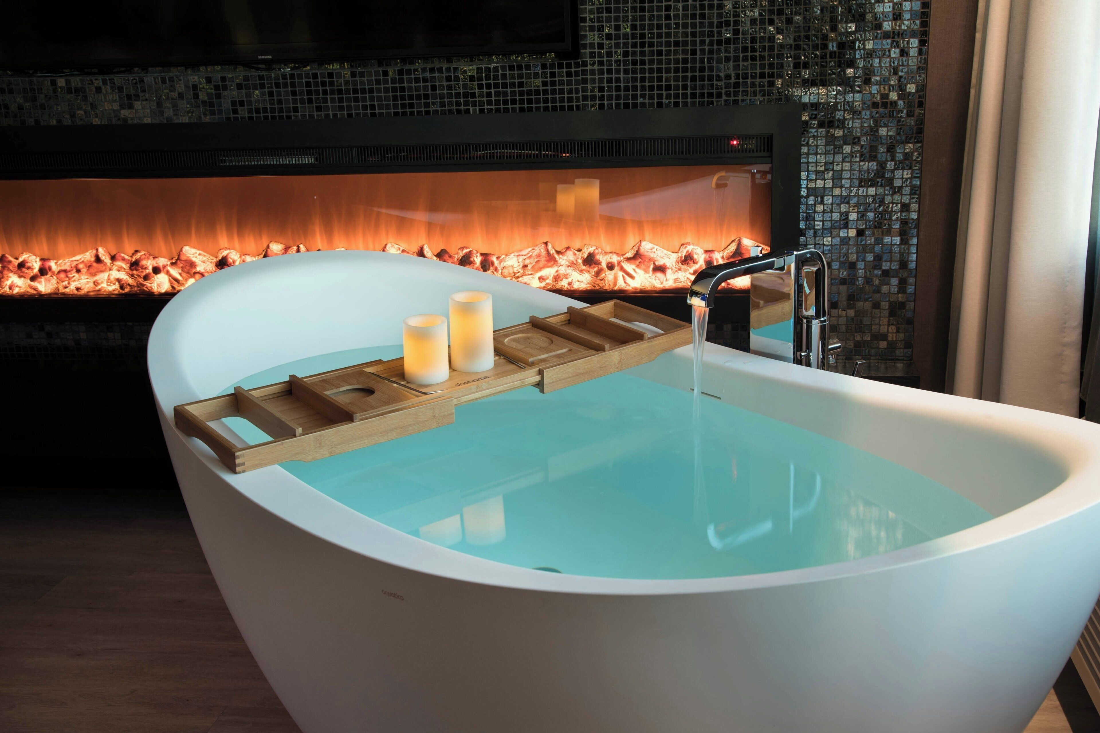 A modern bathroom with a freestanding bathtub, a wooden tray with candles, and a tiled wall with a mosaic design.