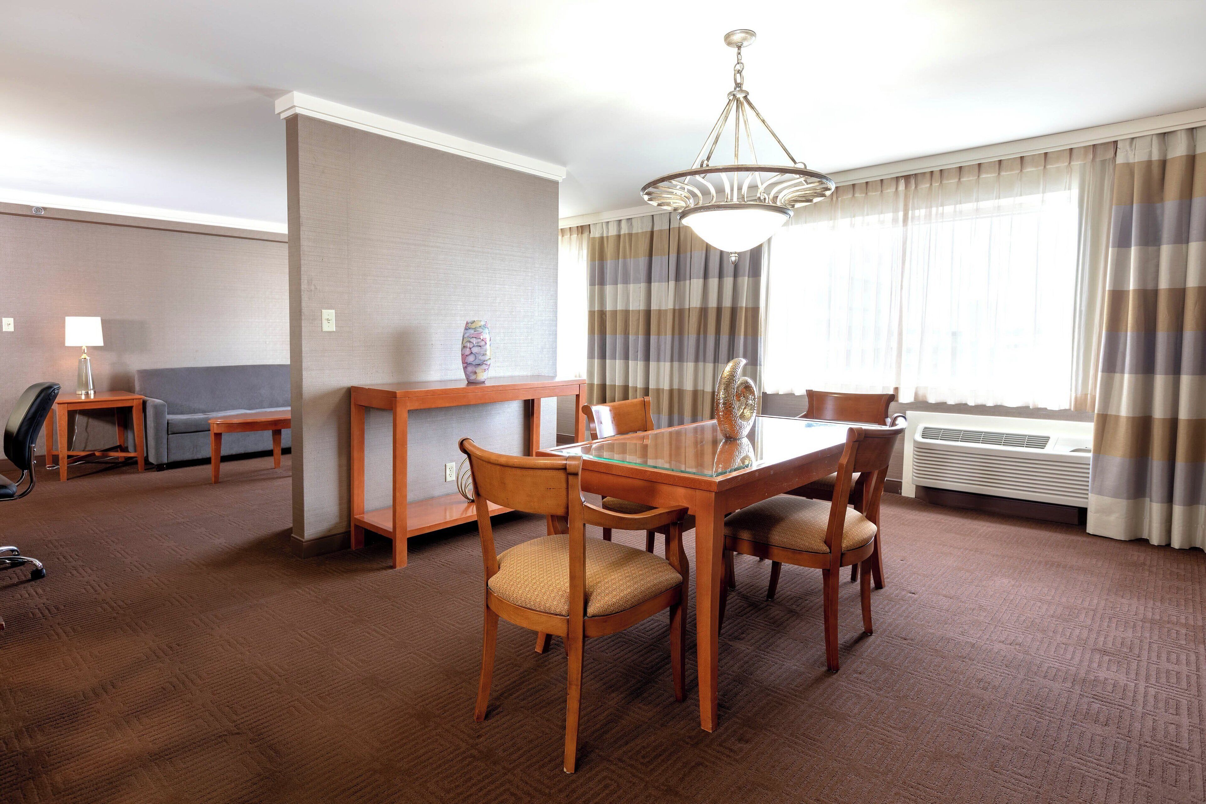 Une chambre d’hôtel dotée d’un coin repas comprenant une table à plateau en verre, des chaises en bois, un lustre, un canapé, une petite table avec une lampe et une fenêtre avec des rideaux.