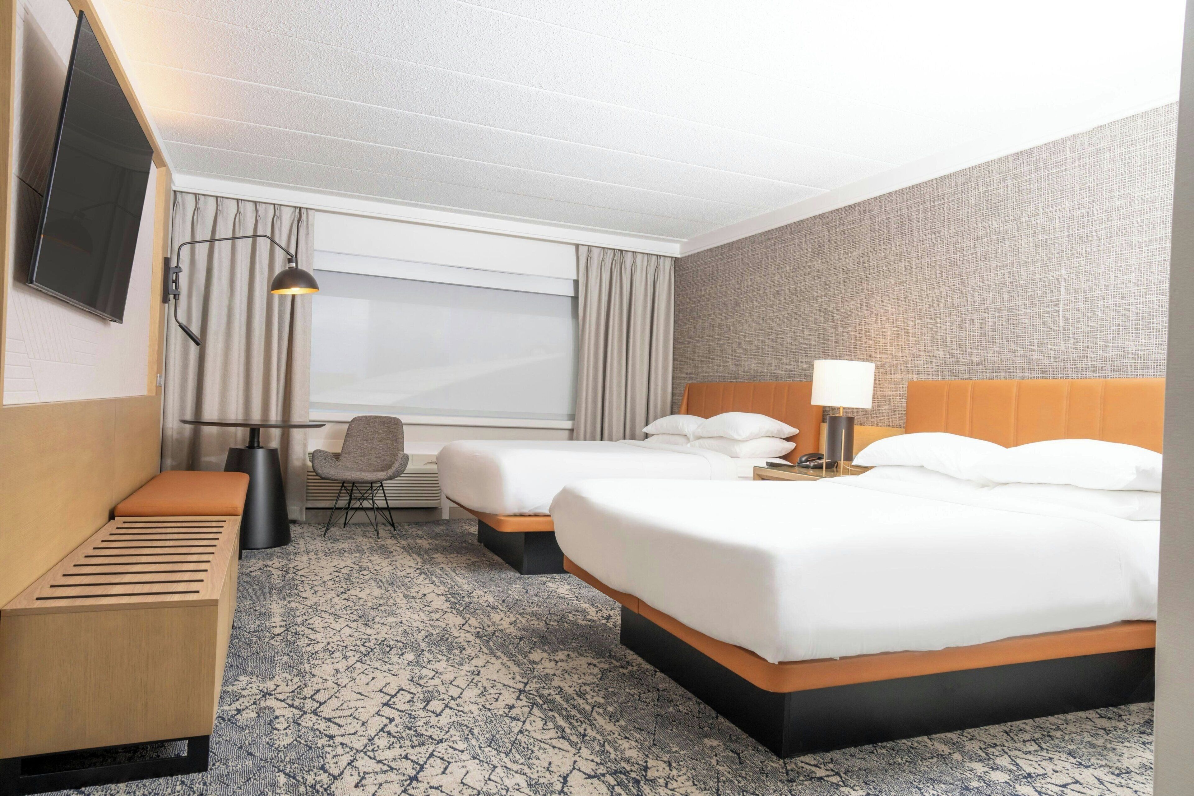 A hotel room with two beds, a desk, a chair, a TV, and a window with curtains.