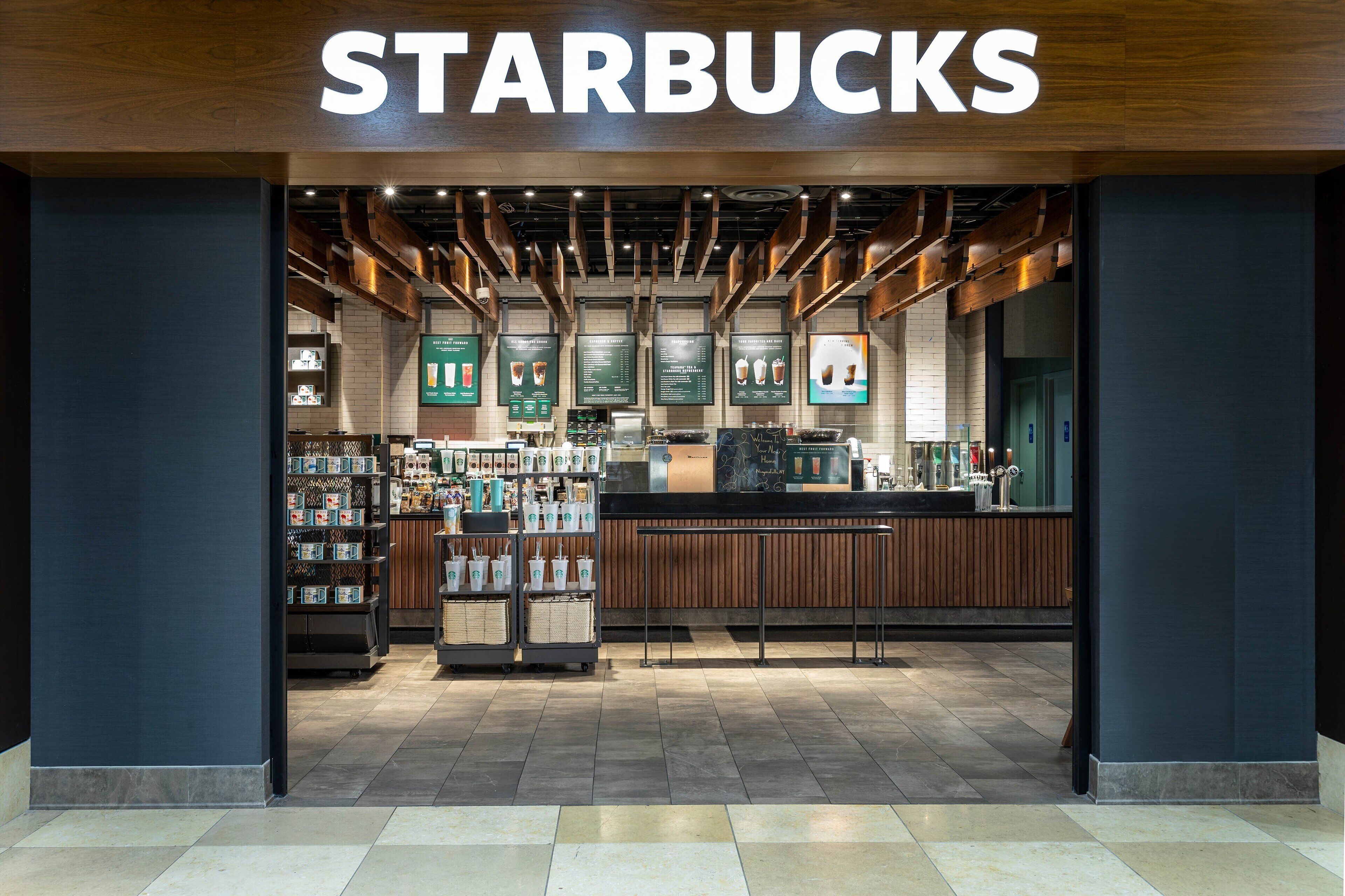 Uma Starbucks com interior moderno, detalhes em madeira e diversas opções de bebidas expostas em prateleiras.