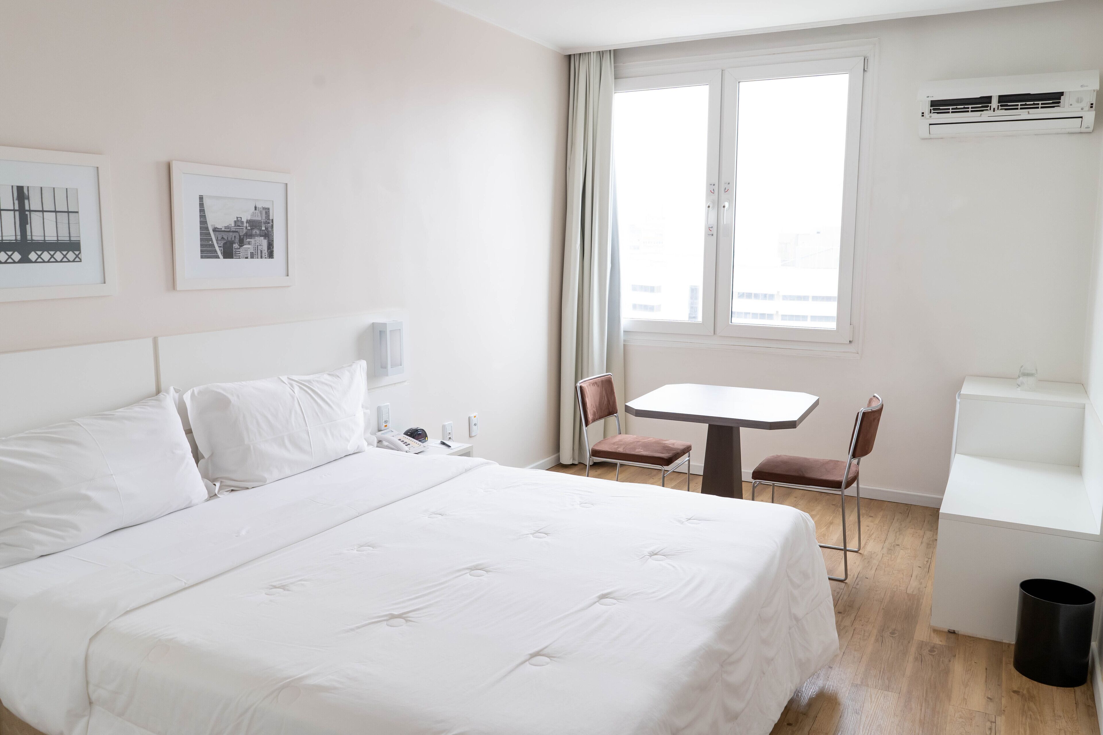 A hotel room with a large bed, a desk, two chairs, a window with curtains, and an air conditioning unit.