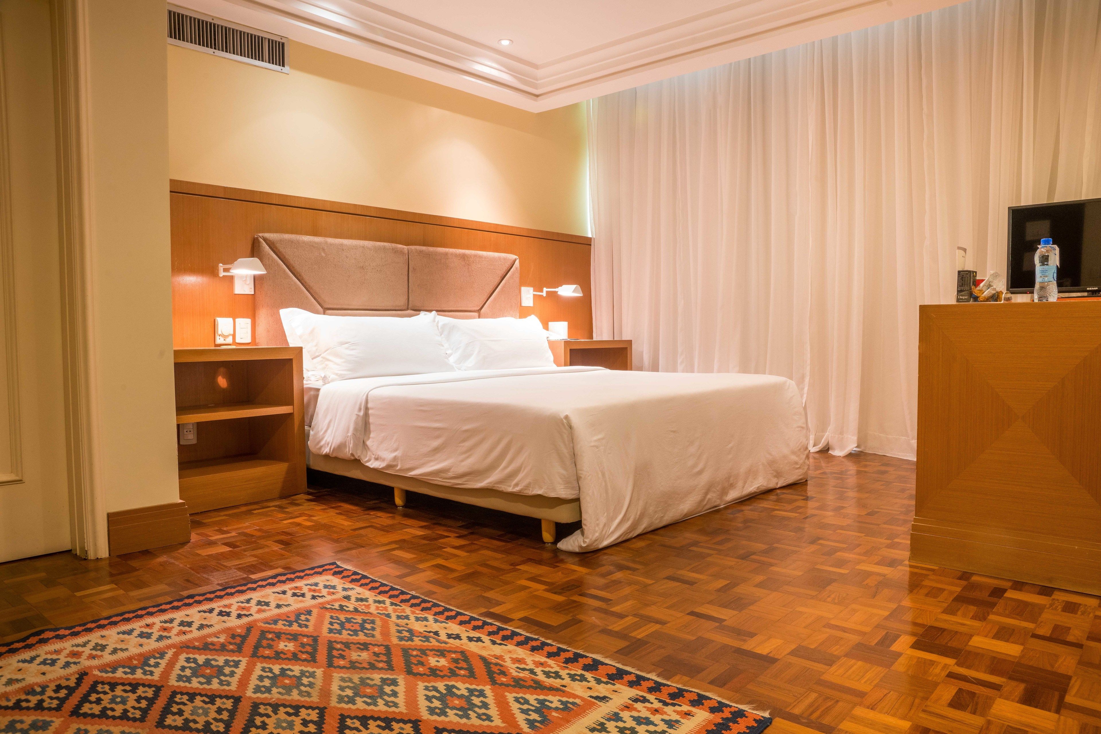 Habitación de hotel con piso de madera, una cama grande con ropa de cama blanca, dos mesitas de noche con lámparas, un televisor sobre un mueble de madera y una alfombra estampada.