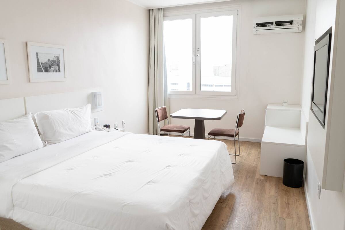 Una camera d'albergo con un letto grande, un tavolino, due sedie, una finestra con tende, un condizionatore d'aria e un cestino.