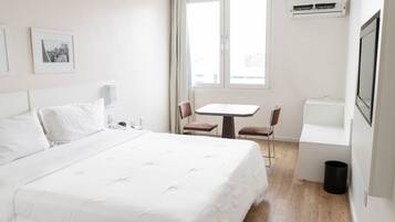Quarto de hotel com uma cama grande, uma mesa pequena, duas cadeiras, uma janela com cortinas, um aparelho de ar condicionado e uma lixeira.
