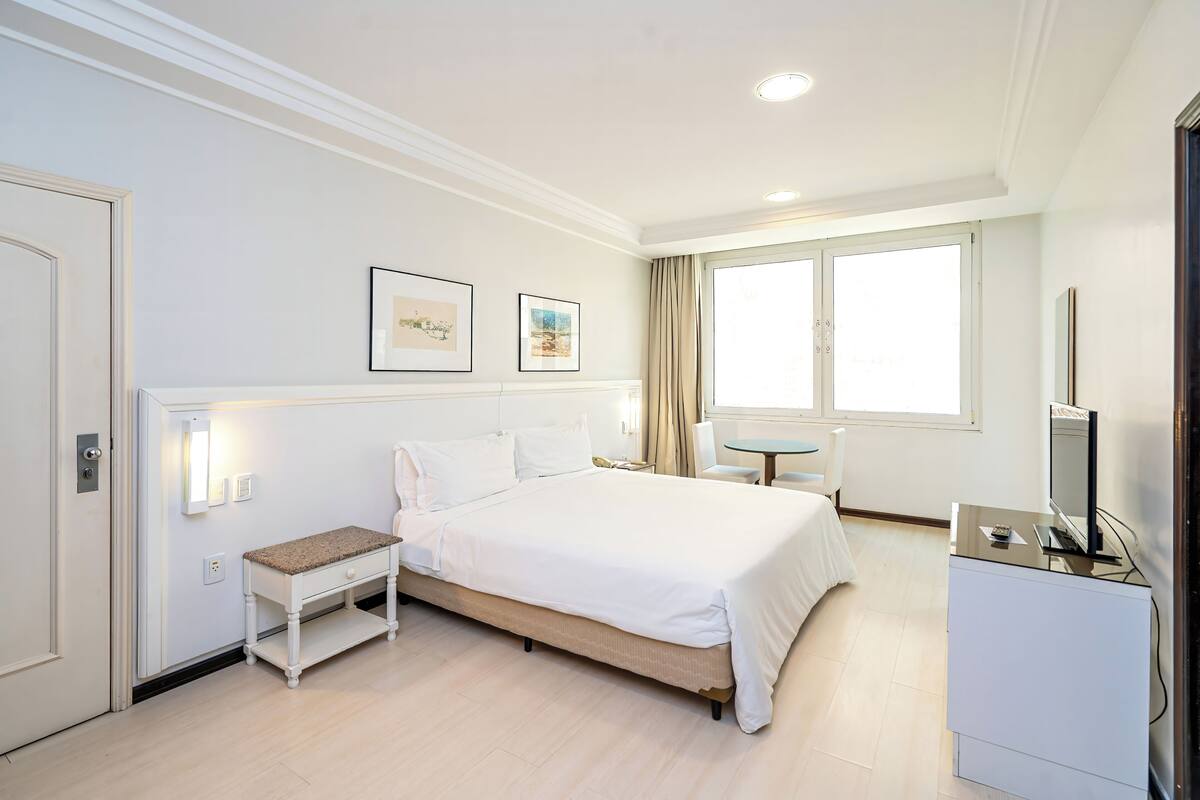 Camera d'albergo con un letto, comodino, sedia, scrivania, televisione e ampie finestre.