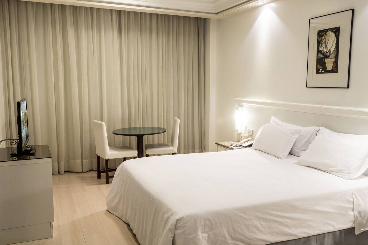Una camera d'albergo con un letto, una scrivania con un computer, una sedia, un tavolino e una finestra con tende.