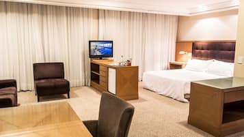 Quarto de hotel com cama, escrivaninha, cadeira, televisão e minibar.