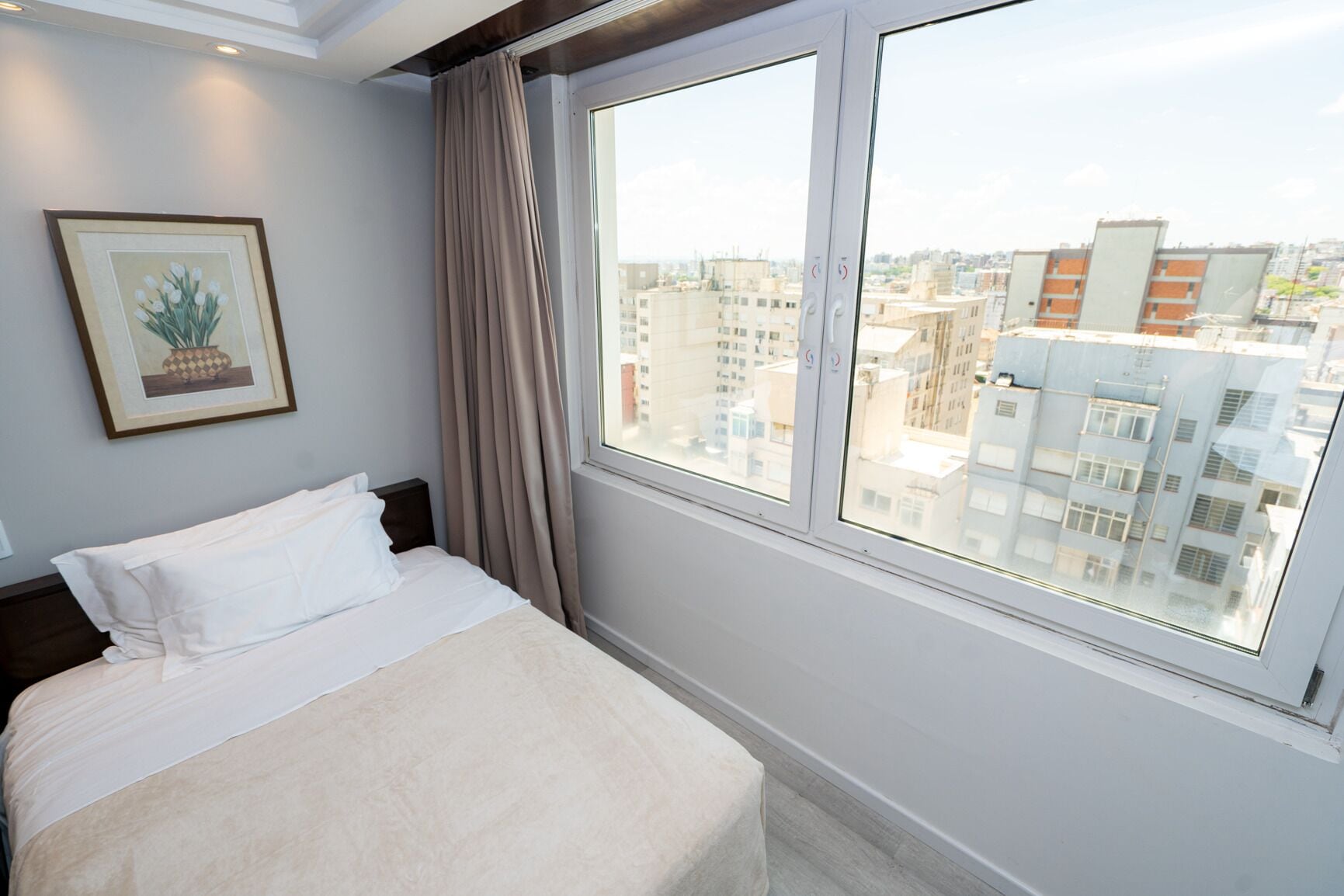 A hotel room with a bed, a window with curtains, a framed picture on the wall, and a view of the cityscape.