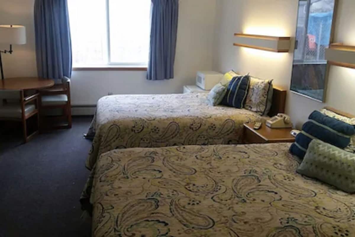 Quarto de hotel com duas camas, uma escrivaninha, uma cadeira, um abajur e uma janela com cortinas.