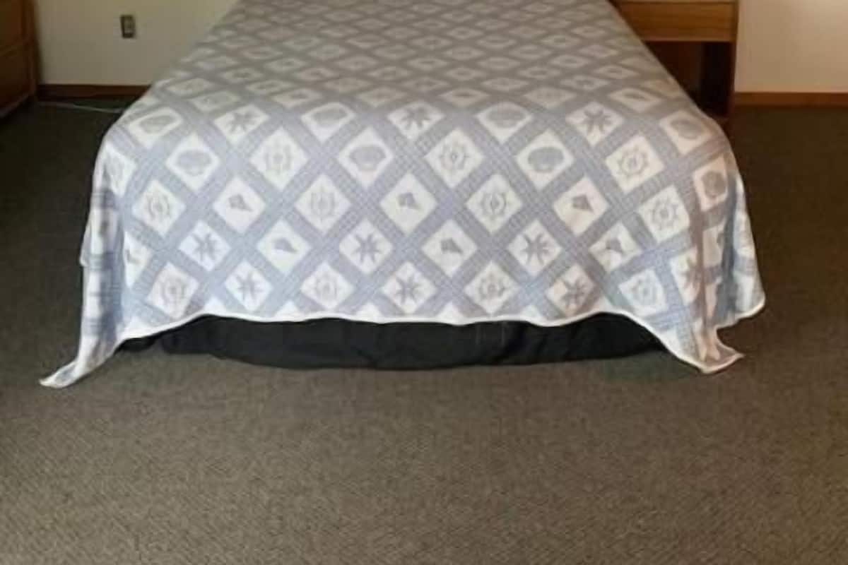 Quarto de hotel com uma cama, mesa de cabeceira com abajur, quadro na parede e luz no teto.