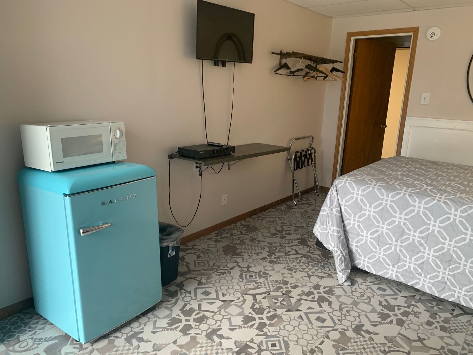 Une chambre d’hôtel avec un réfrigérateur bleu, un four à micro-ondes, un téléviseur à écran plat fixé au mur, un lit avec une literie à motifs et une petite étagère sur laquelle sont posés quelques objets.