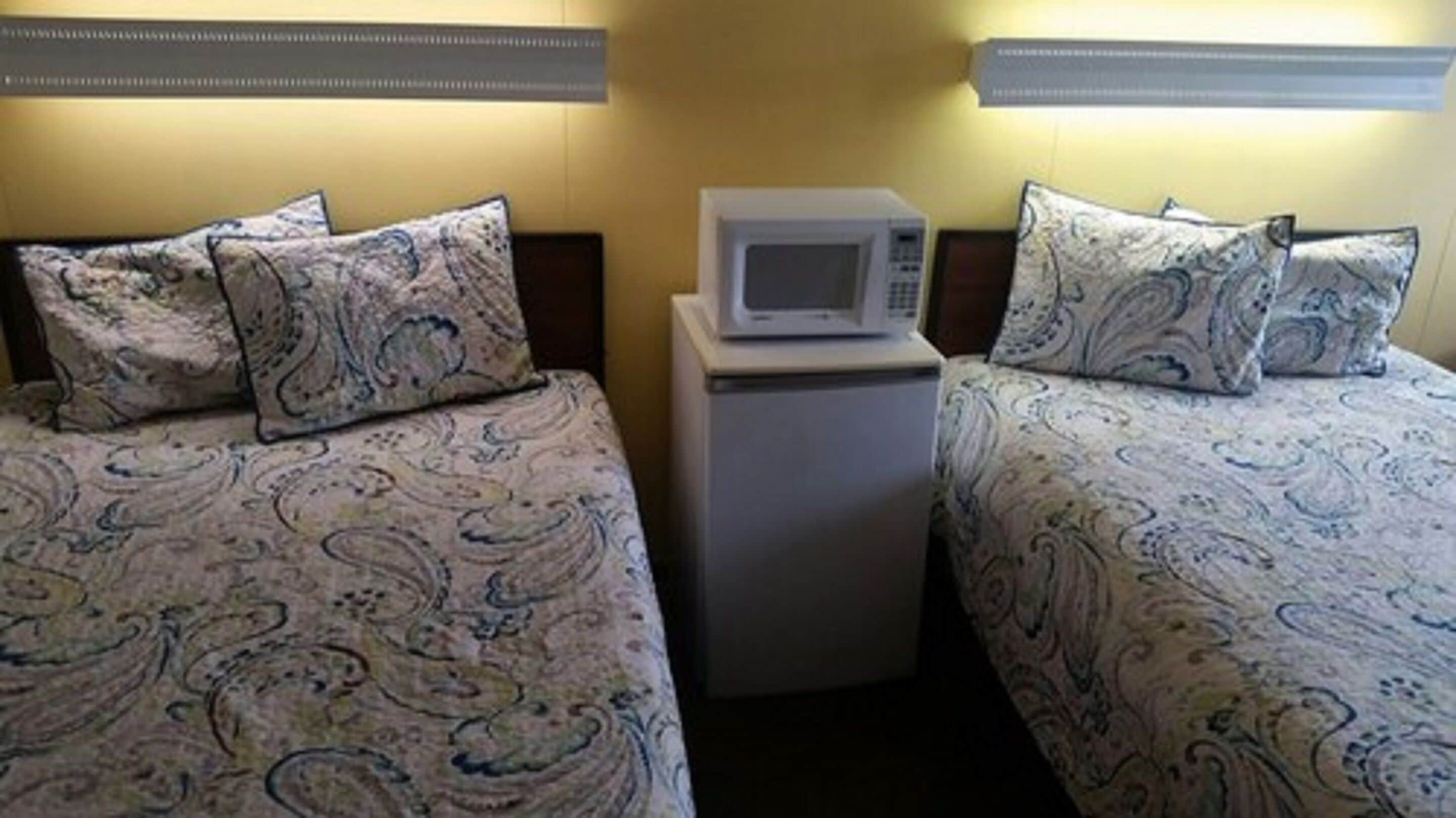 A hotel room with two beds, each with patterned bedding and pillows, and a small white microwave on a table between the beds.