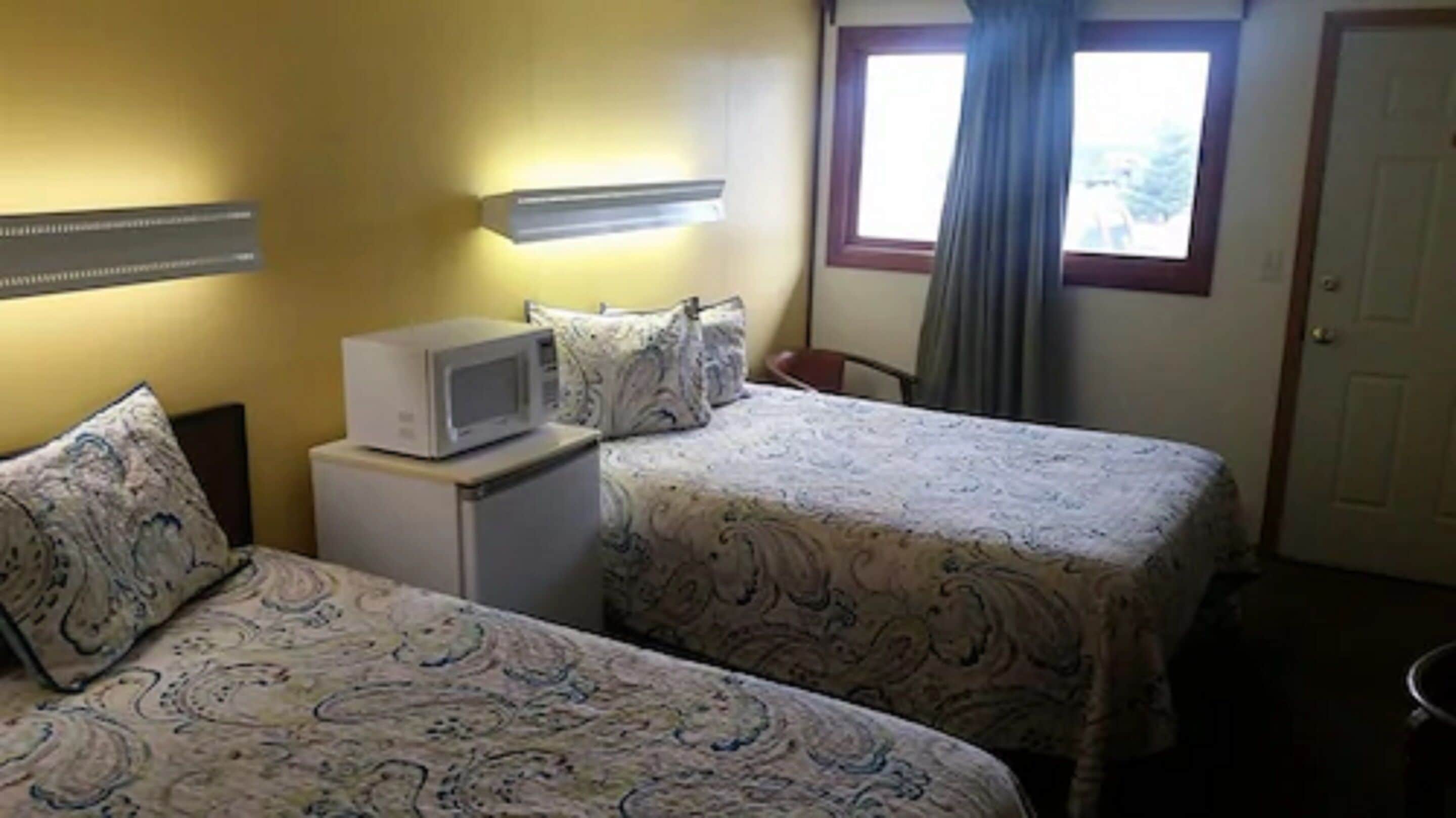 A hotel room with two beds, a microwave on a small table, a window with curtains, and a door.