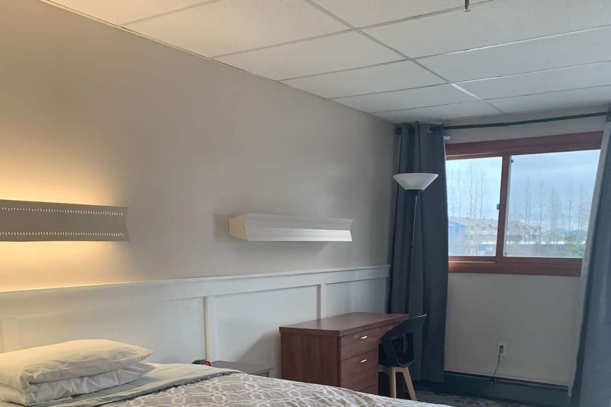 Quarto de hotel com uma cama grande, uma escrivaninha, uma cadeira, um abajur e uma janela com vista para árvores.