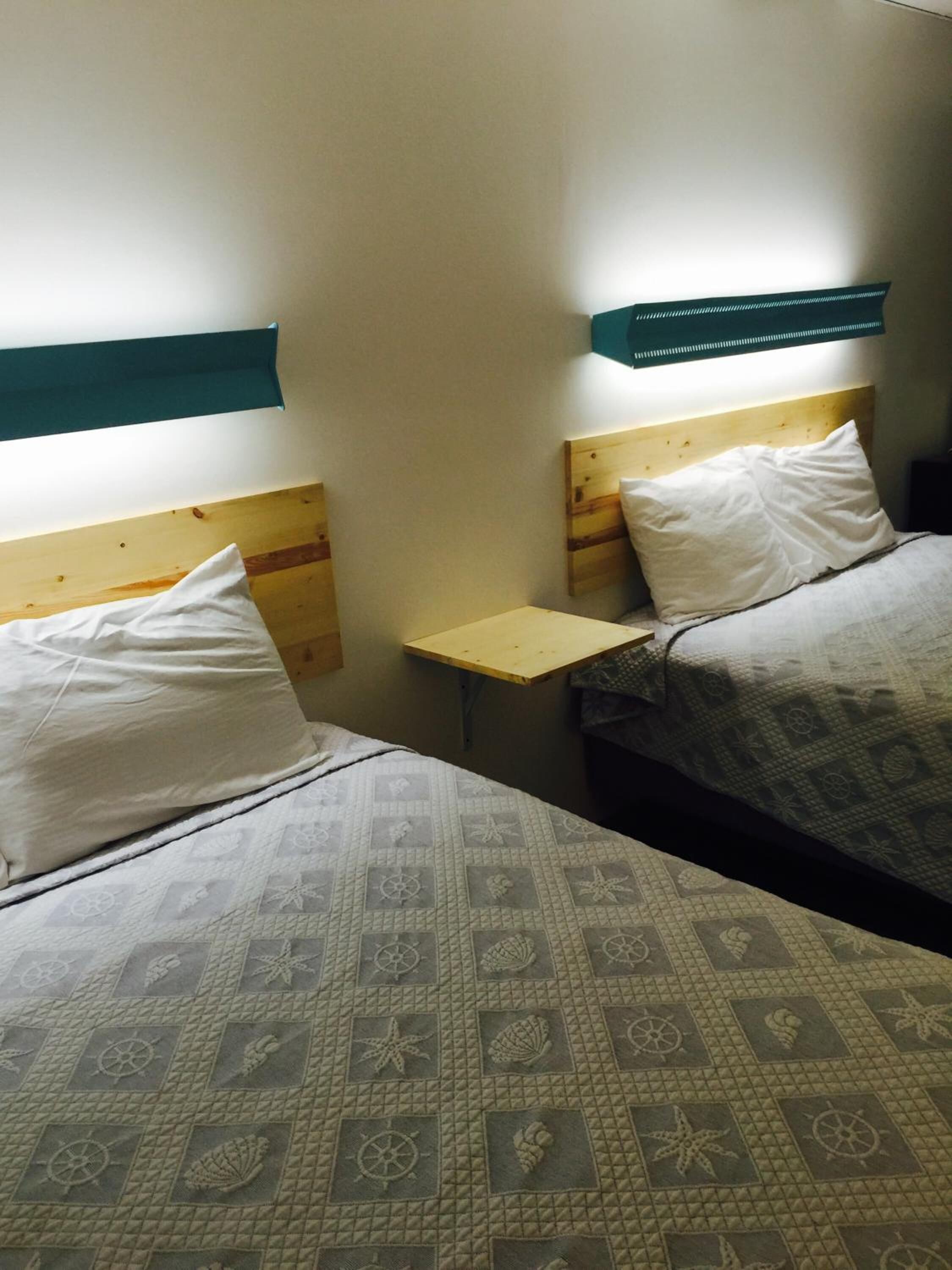 A small, simple room with two beds, a small table, and a headboard with a light fixture.