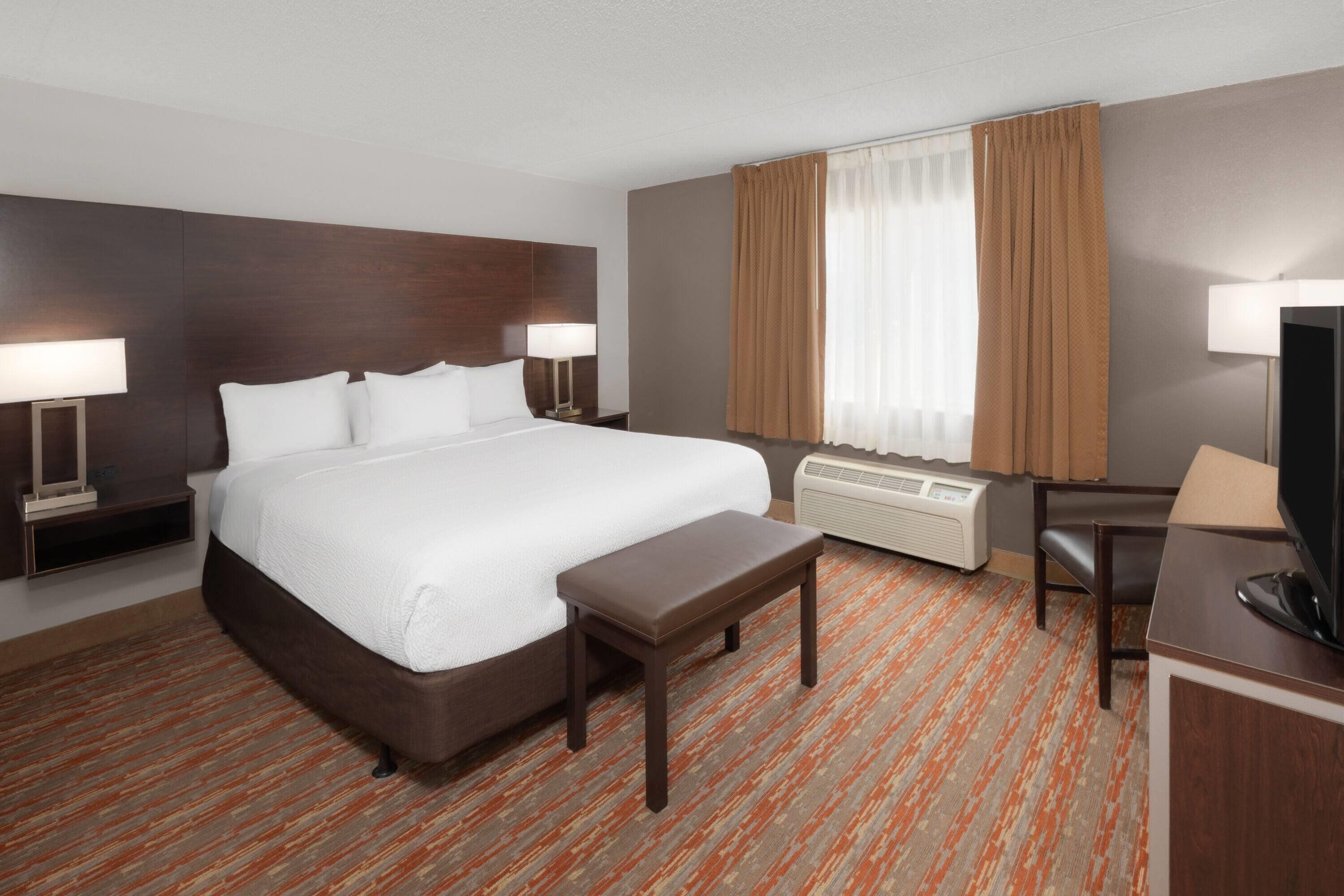 Ein Hotelzimmer mit einem großen Bett, Nachttischen, einem Schreibtisch, einem Sessel und einem Fernseher.