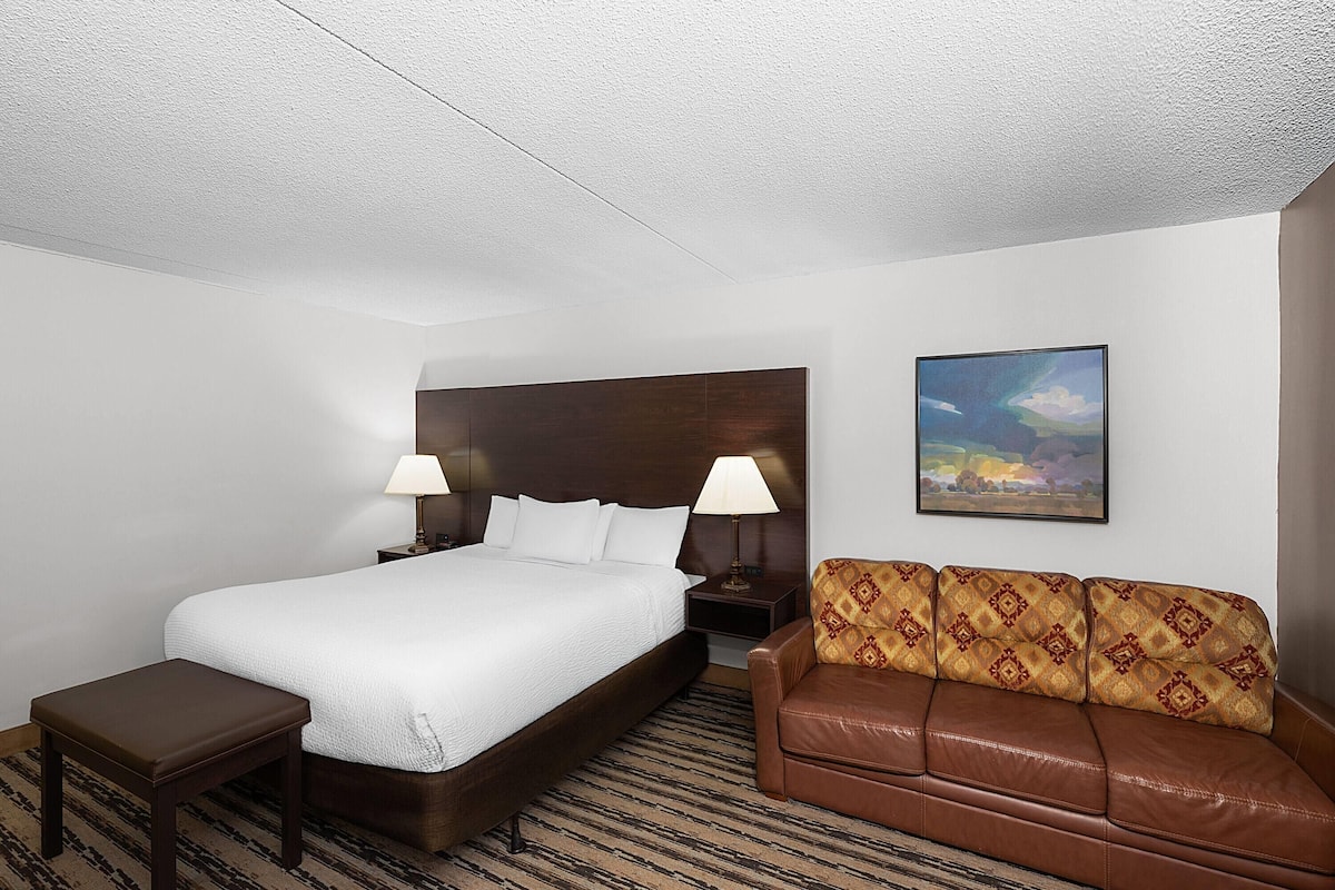 A hotel room with a large bed, two bedside tables with lamps, a brown leather sofa, and a painting on the wall.