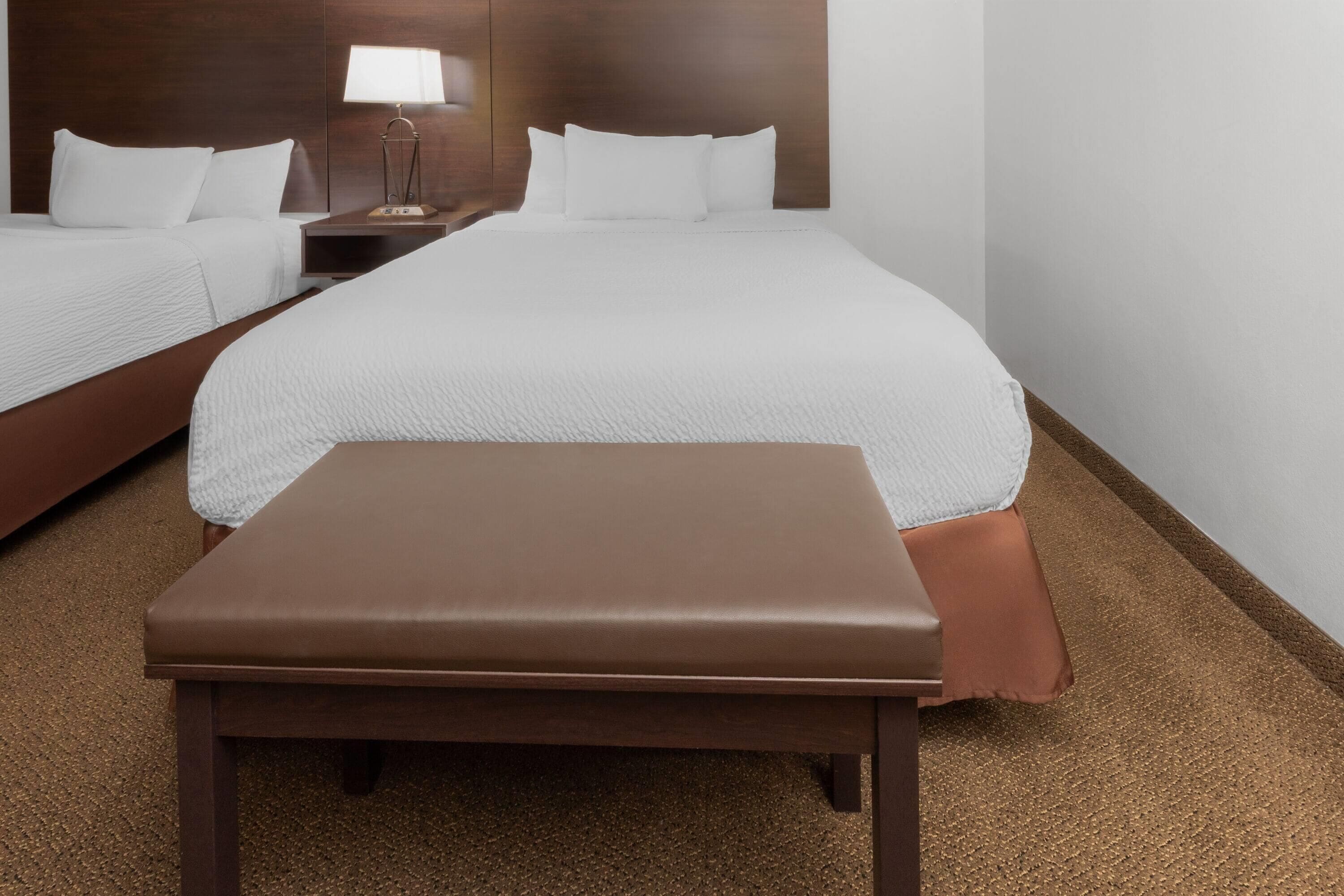 A hotel room with two beds, a bench, and a nightstand with a lamp.