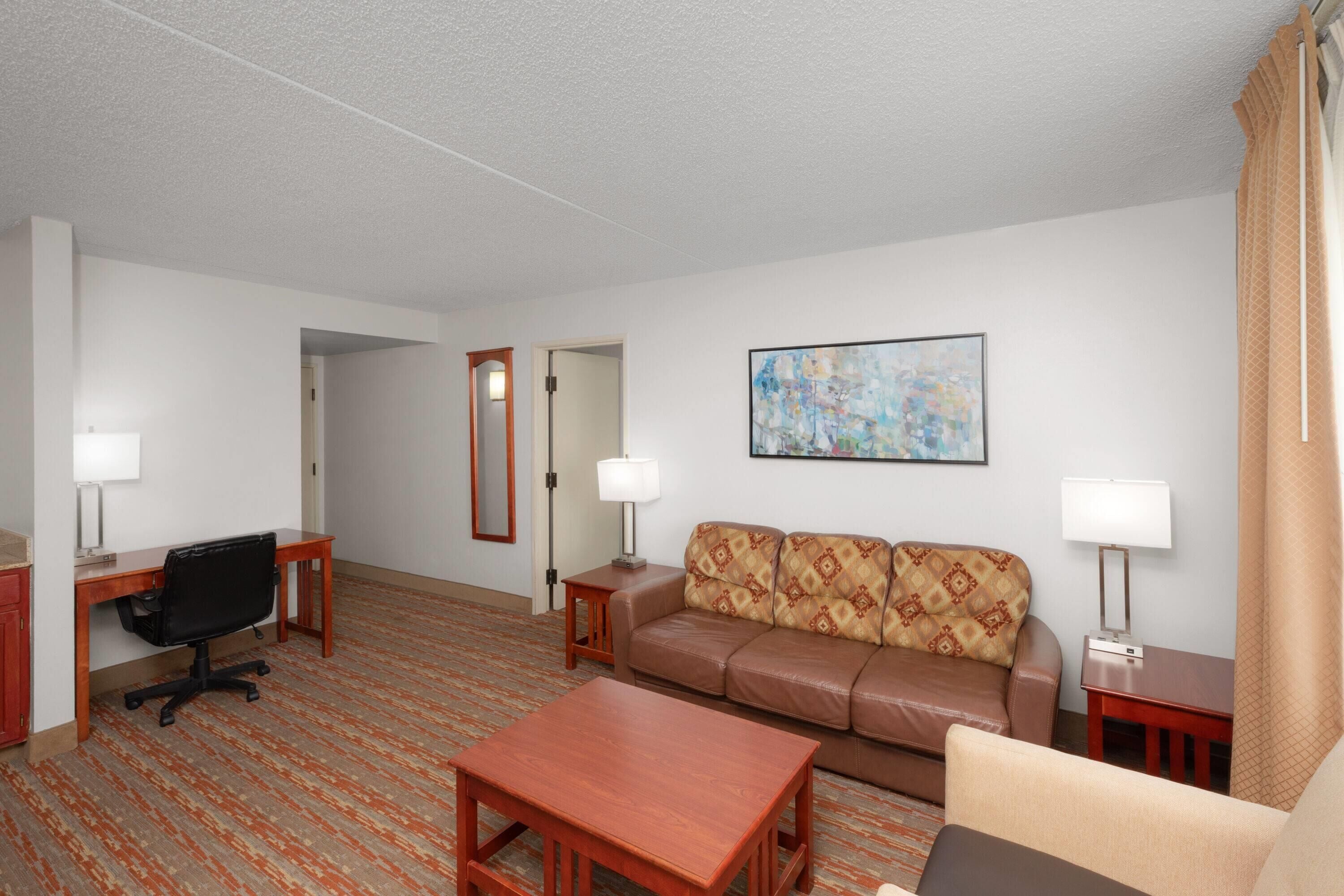 A hotel room with a sofa, a desk with a chair, a coffee table, a lamp, and a painting on the wall.