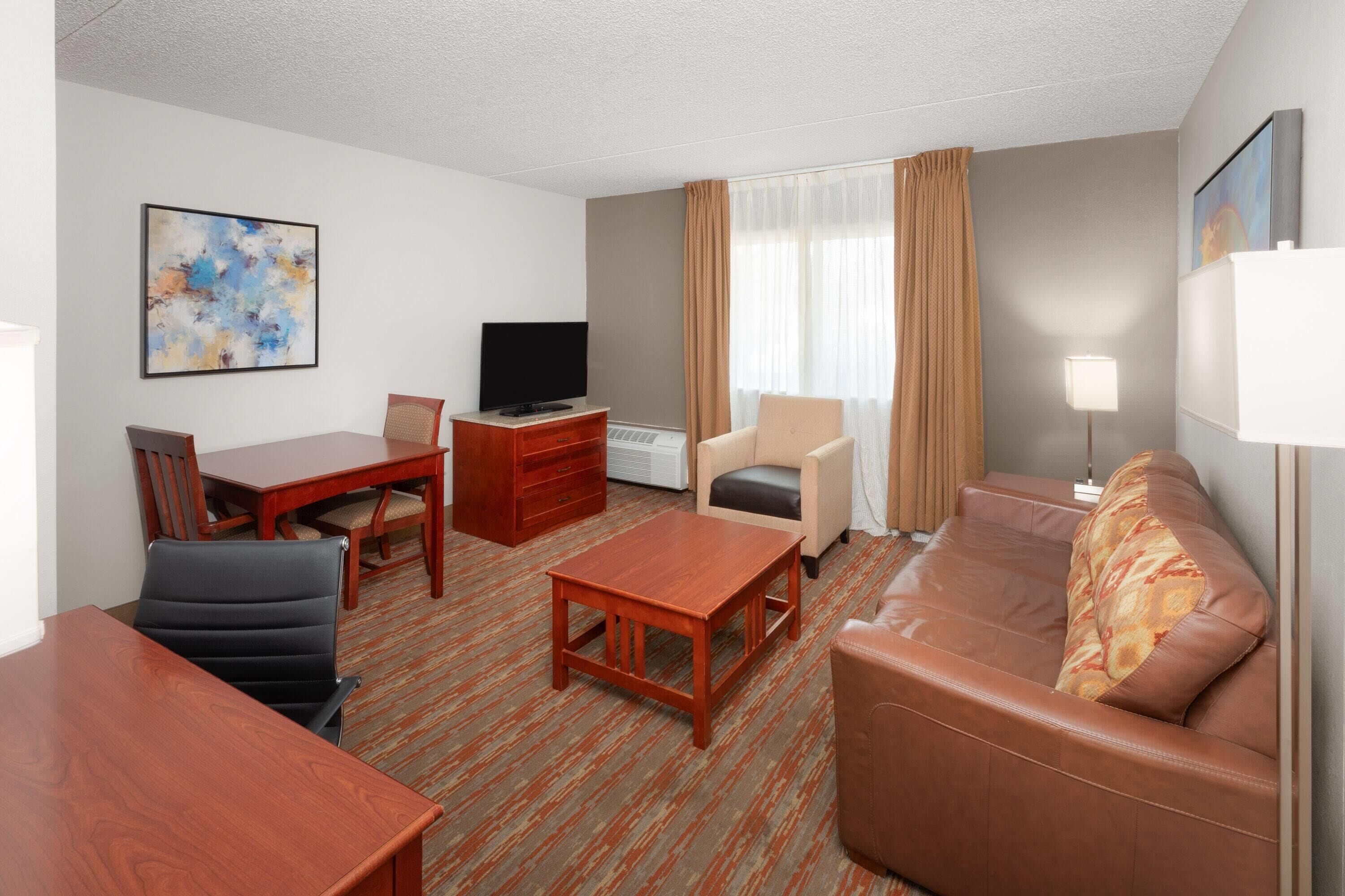 A hotel room with a wooden dining table, a sofa, a TV, and a lamp.