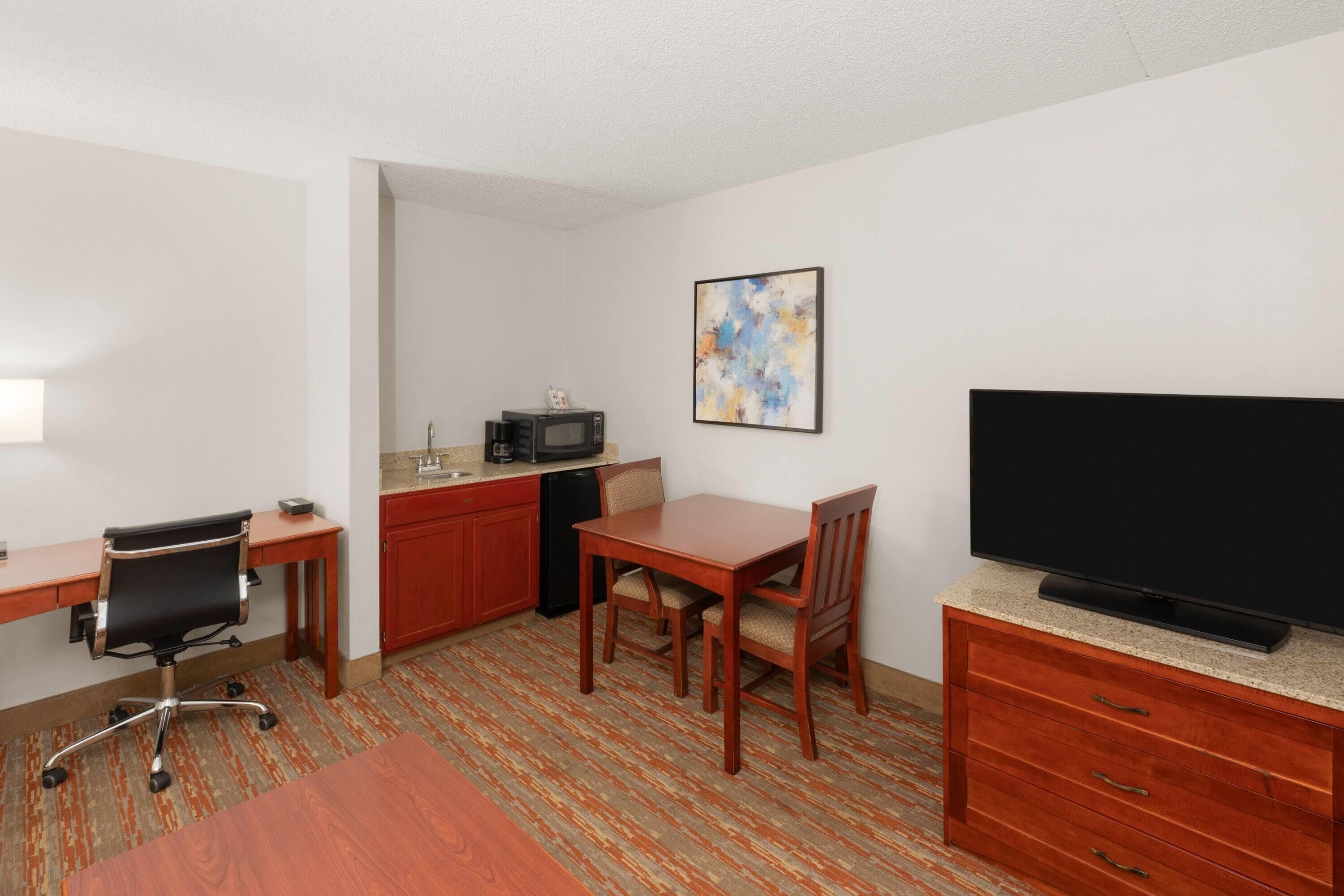 Ein Hotelzimmer mit Holzschreibtisch, einem Esstisch, einem Fernseher auf einem Schrank, einer Mikrowelle und einer kleinen Küchenzeile.