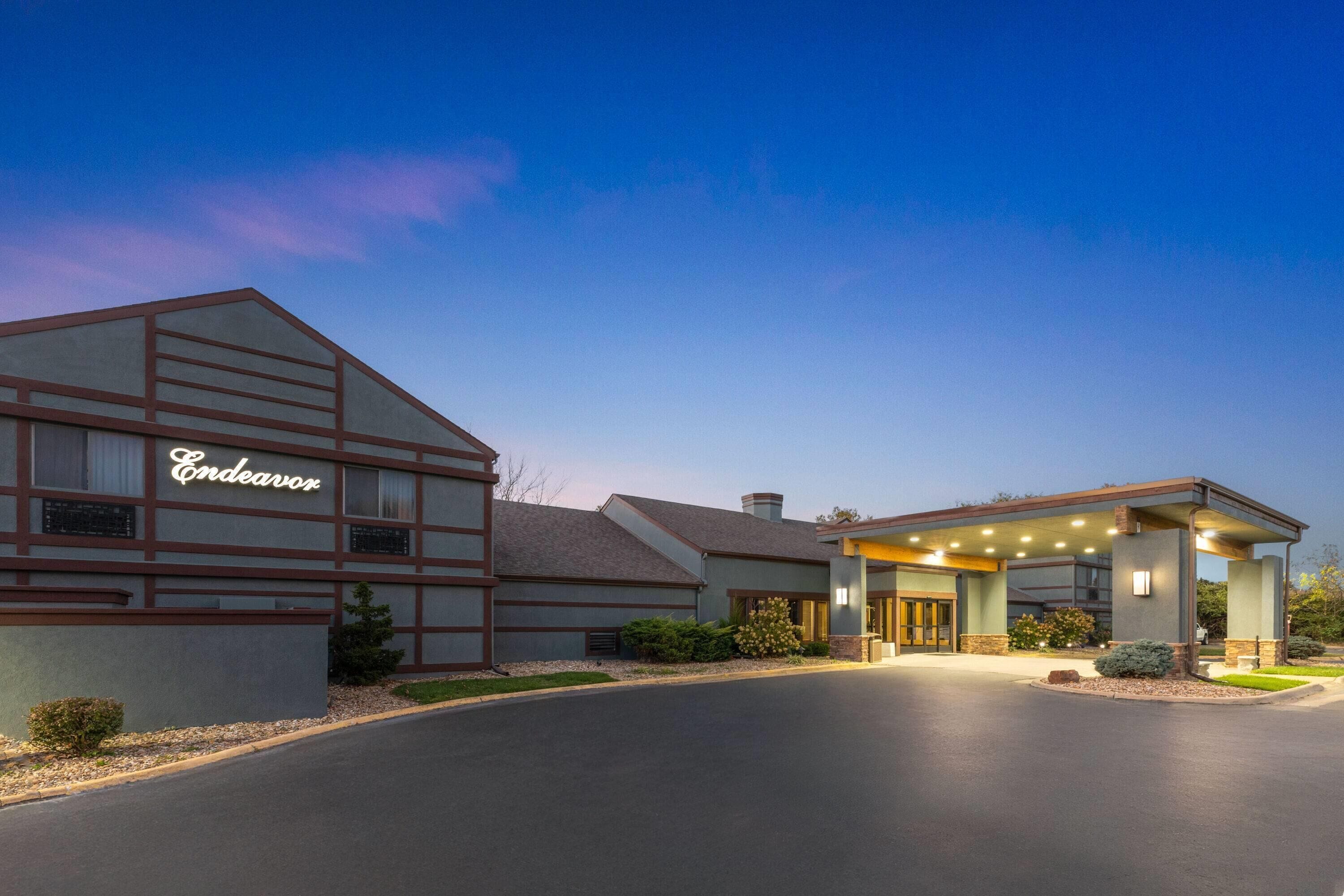 Um hotel com design moderno, exibindo uma placa grande com a inscrição 'Endeavor', uma entrada coberta e um acesso por uma estrada bem cuidada.
