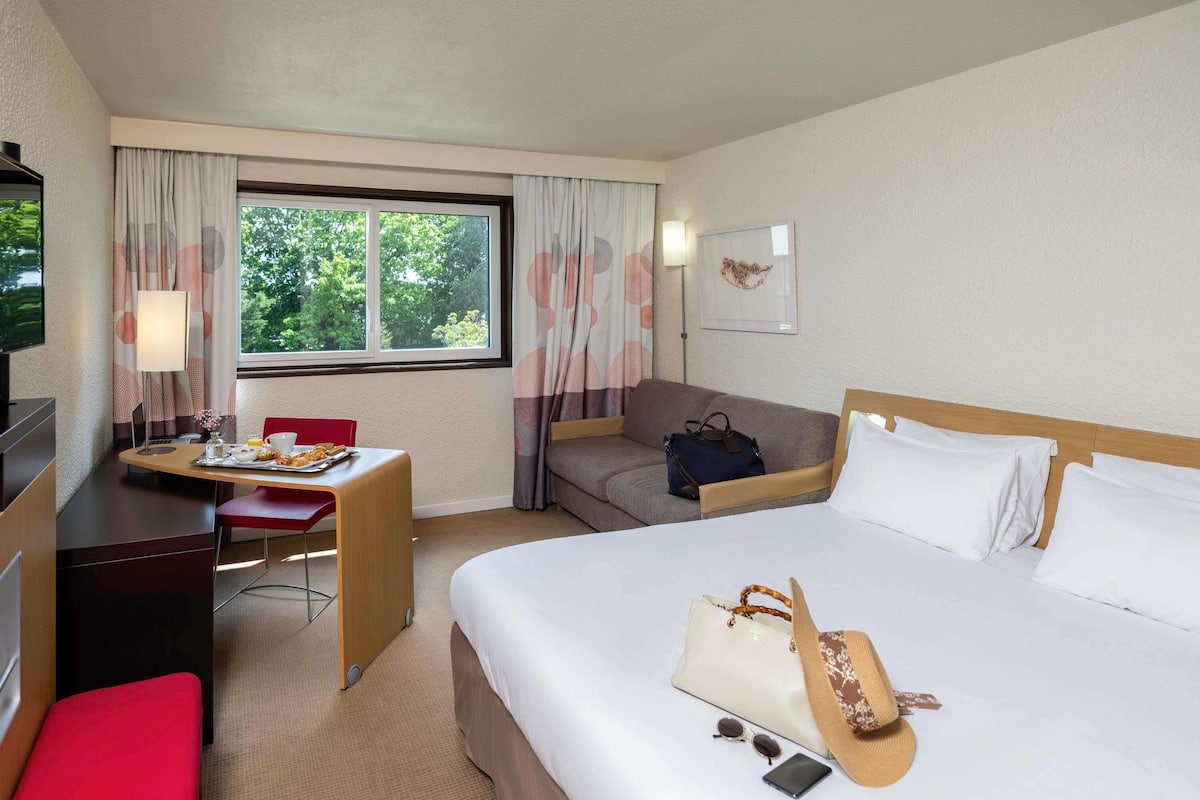 Ein Hotelzimmer mit Bett, Schreibtisch, Stuhl, Sofa, Fernseher und einem Fenster mit Vorhängen.