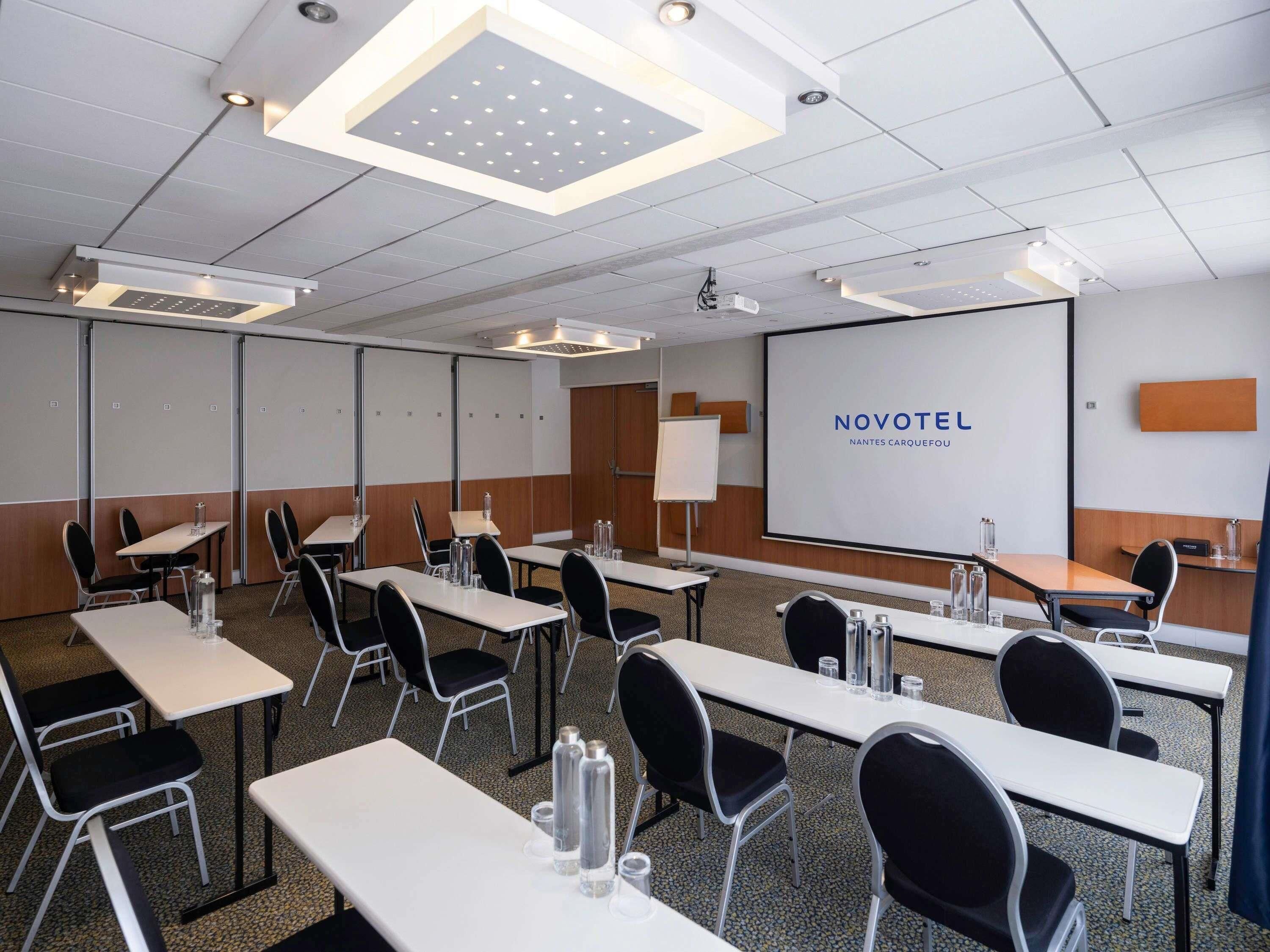 Une salle de conférence équipée de tables, de chaises, d’un écran de projection et d’une présentation intitulée « NOVOTEL NANTES CARQUEFOU ».