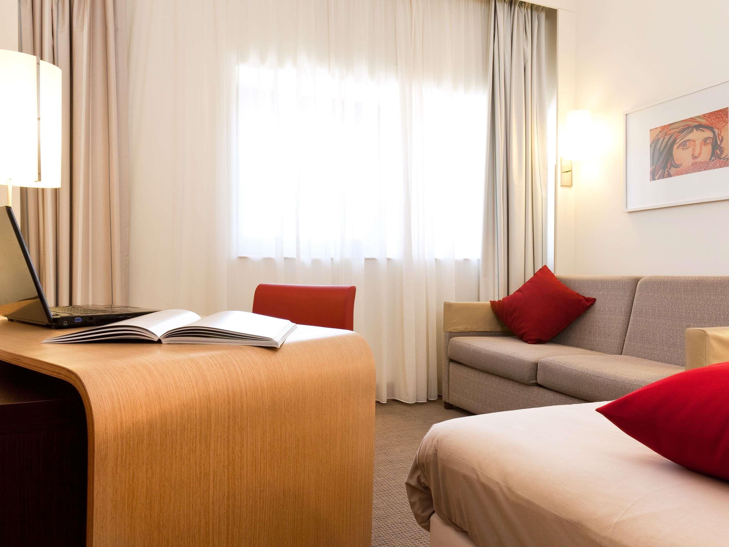 Habitación de hotel con escritorio, computadora portátil, libro abierto, dos sofás y una cama.