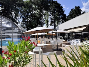 Zona de piscina al aire libre con sillas y sombrillas blancas, rodeada de árboles y una estructura de vidrio.