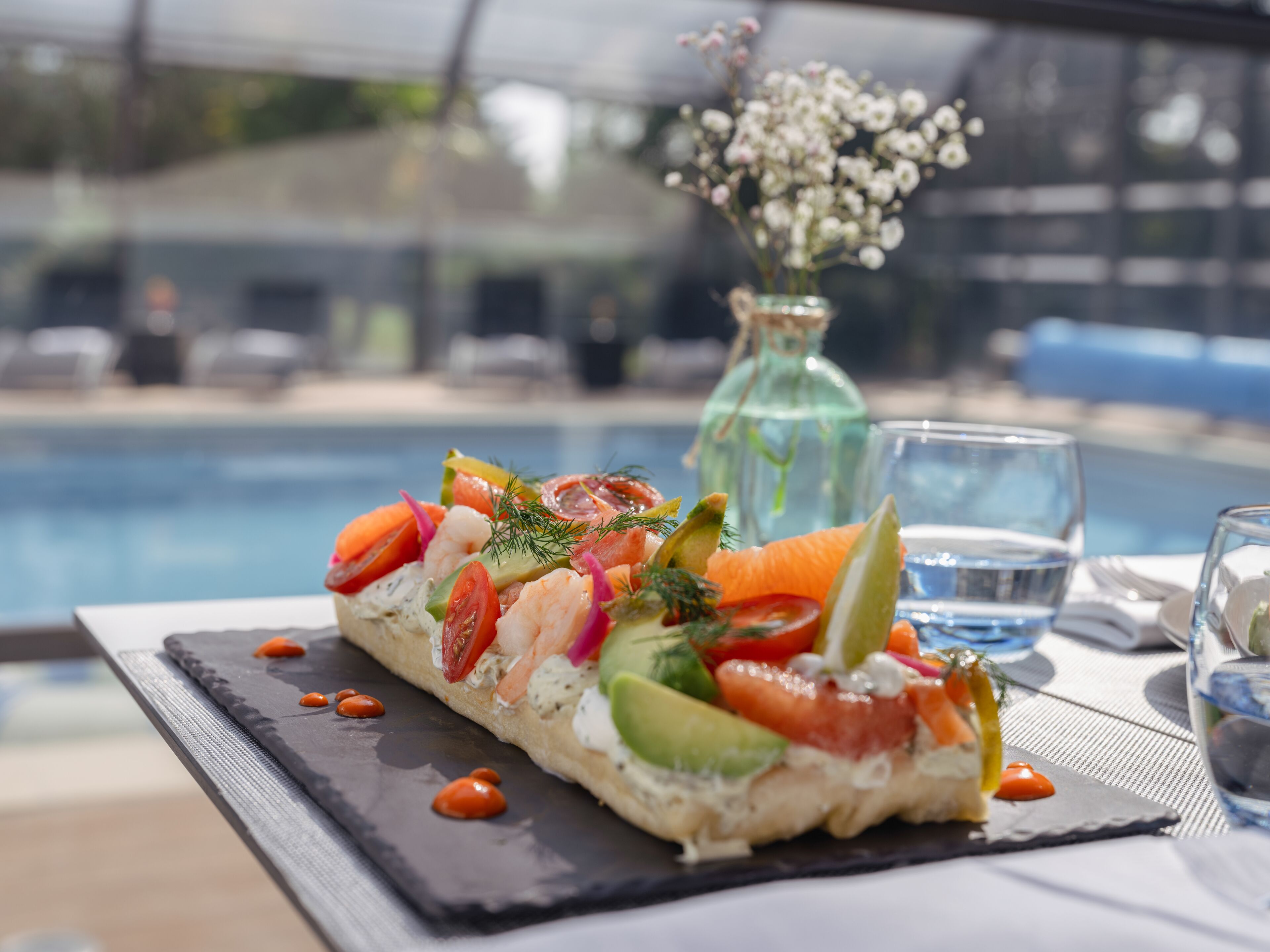 Uma entrada de frutos do mar sobre uma tábua de ardósia, ao lado de uma mesa à beira da piscina, com um copo de água e um vaso de flores.