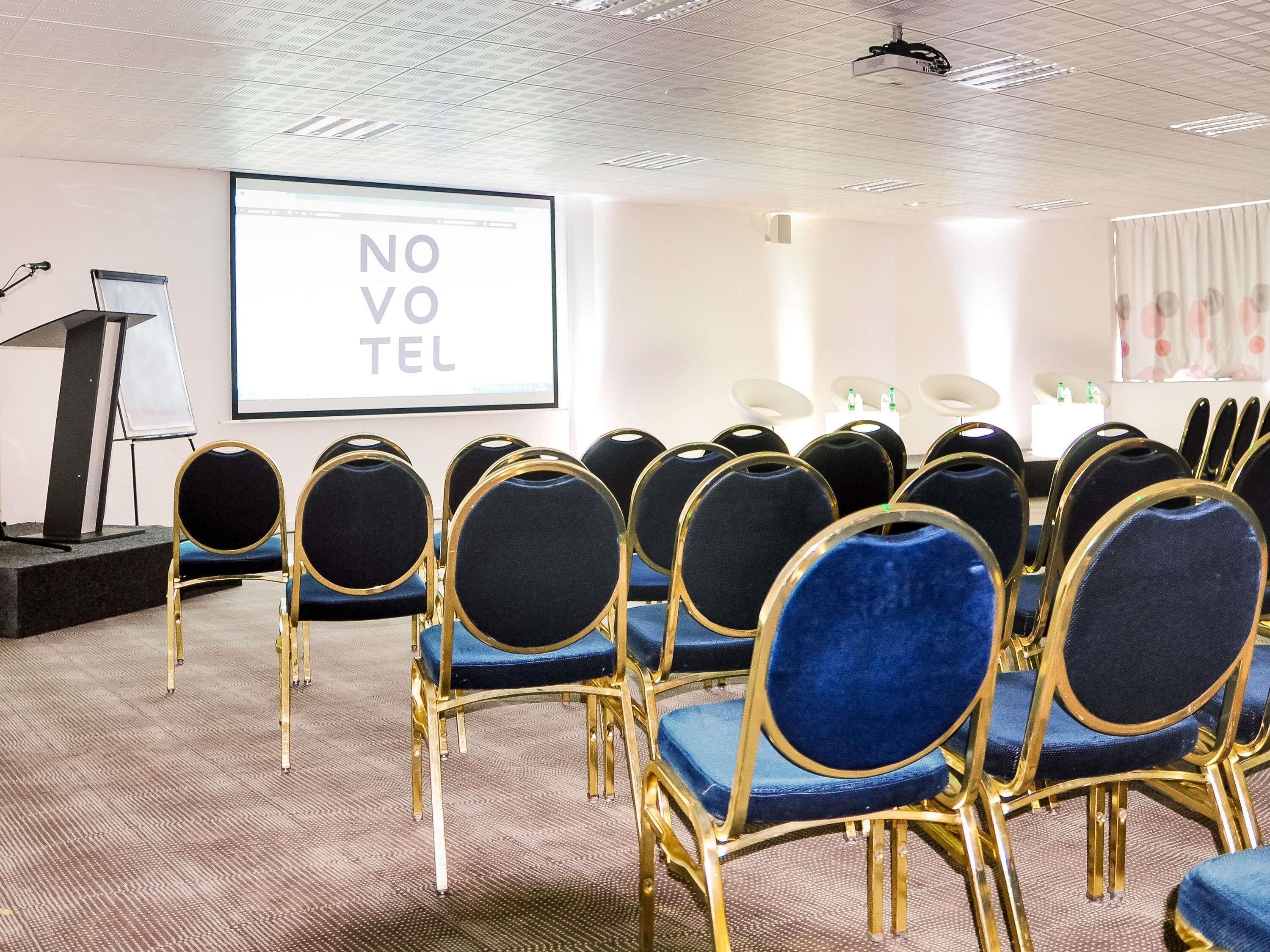 A conference room with rows of blue chairs, a podium, and a screen displaying 'NOVO VEL'.