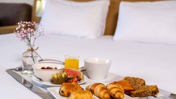 A breakfast tray with croissants, a fruit salad, a glass of orange juice, and a bowl of cereal on a hotel bed.