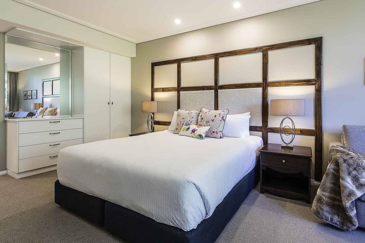 Une chambre à coucher comprenant un grand lit, une commode, une table de chevet, un miroir et une tête de lit dotée d’un luminaire.