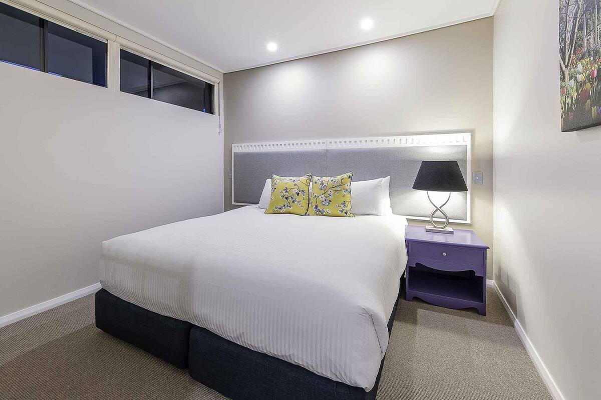 Un dormitorio con cama, mesita de noche, lámpara y ventana.