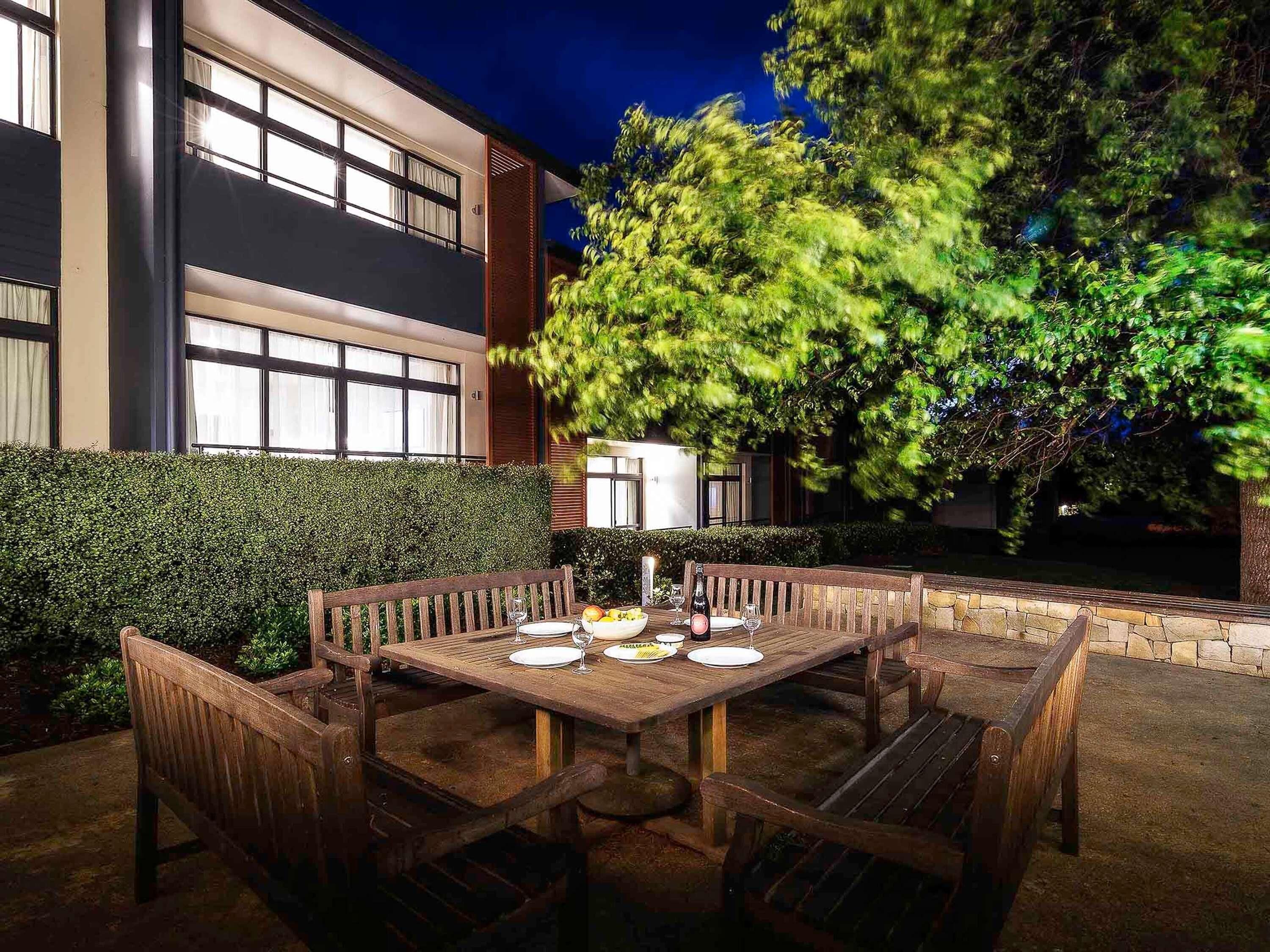 An outdoor dining area with a table set for four, surrounded by wooden chairs, in front of a building with large windows and a hedge.