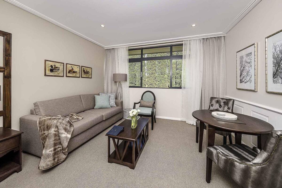 A living room with a sofa, coffee table, and dining table with chairs. There are framed pictures on the walls and a window with curtains.