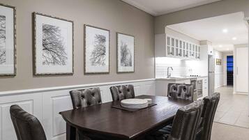 A dining area with a dark wooden table and leather chairs, framed artwork on the wall, and a kitchen in the background.