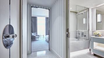 Quarto de hotel moderno com um espelho grande, box de vidro para o chuveiro e vista para o quarto vizinho.