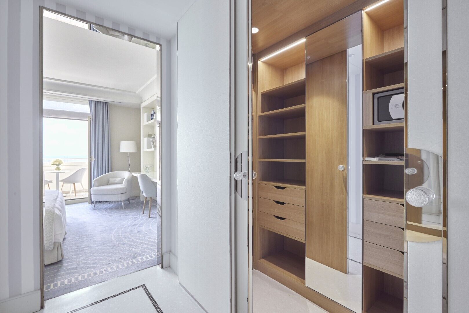 A hotel room with a walk-in closet, a sitting area with a chair and a table, and a view of the beach through the window.