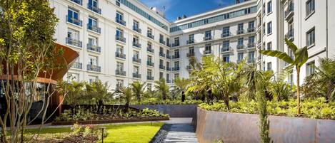 A modern hotel courtyard with a central pathway, surrounded by lush greenery and a white building with multiple balconies.