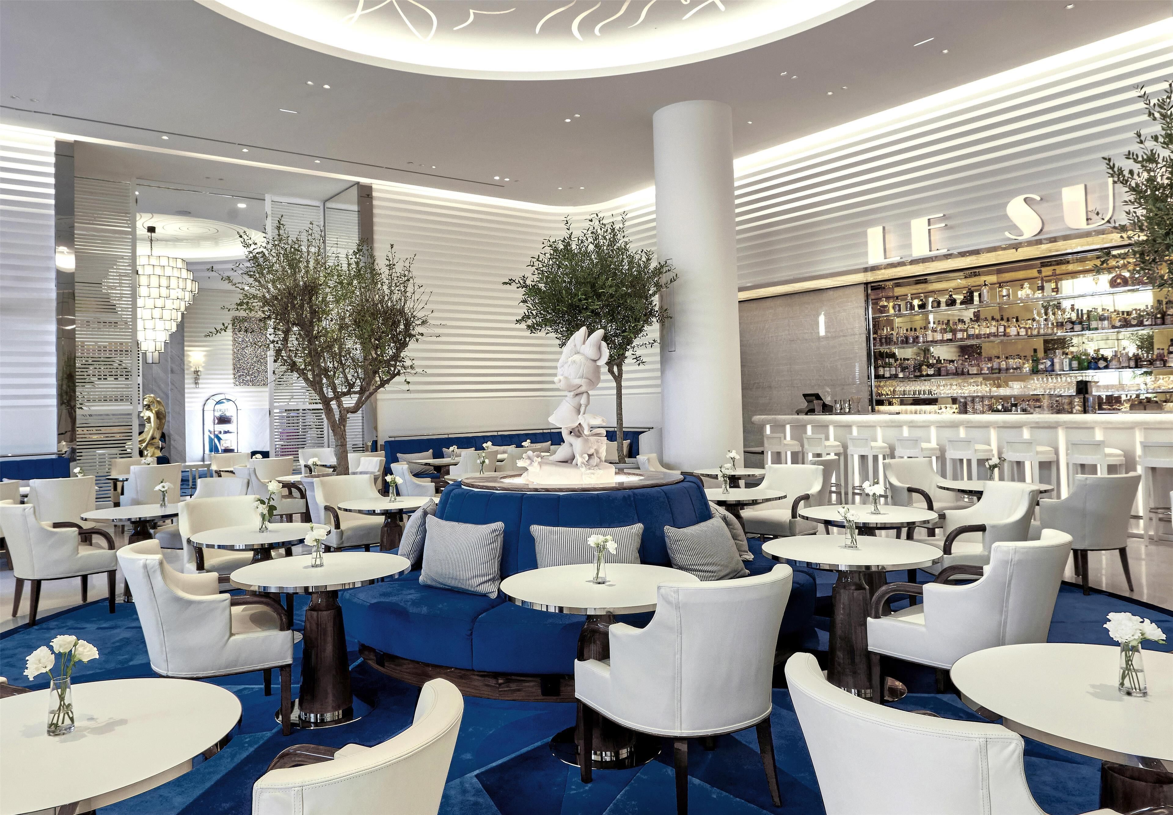 Un área de bar moderna con mesas redondas y sillas blancas, un sofá azul y un letrero iluminado grande que dice 