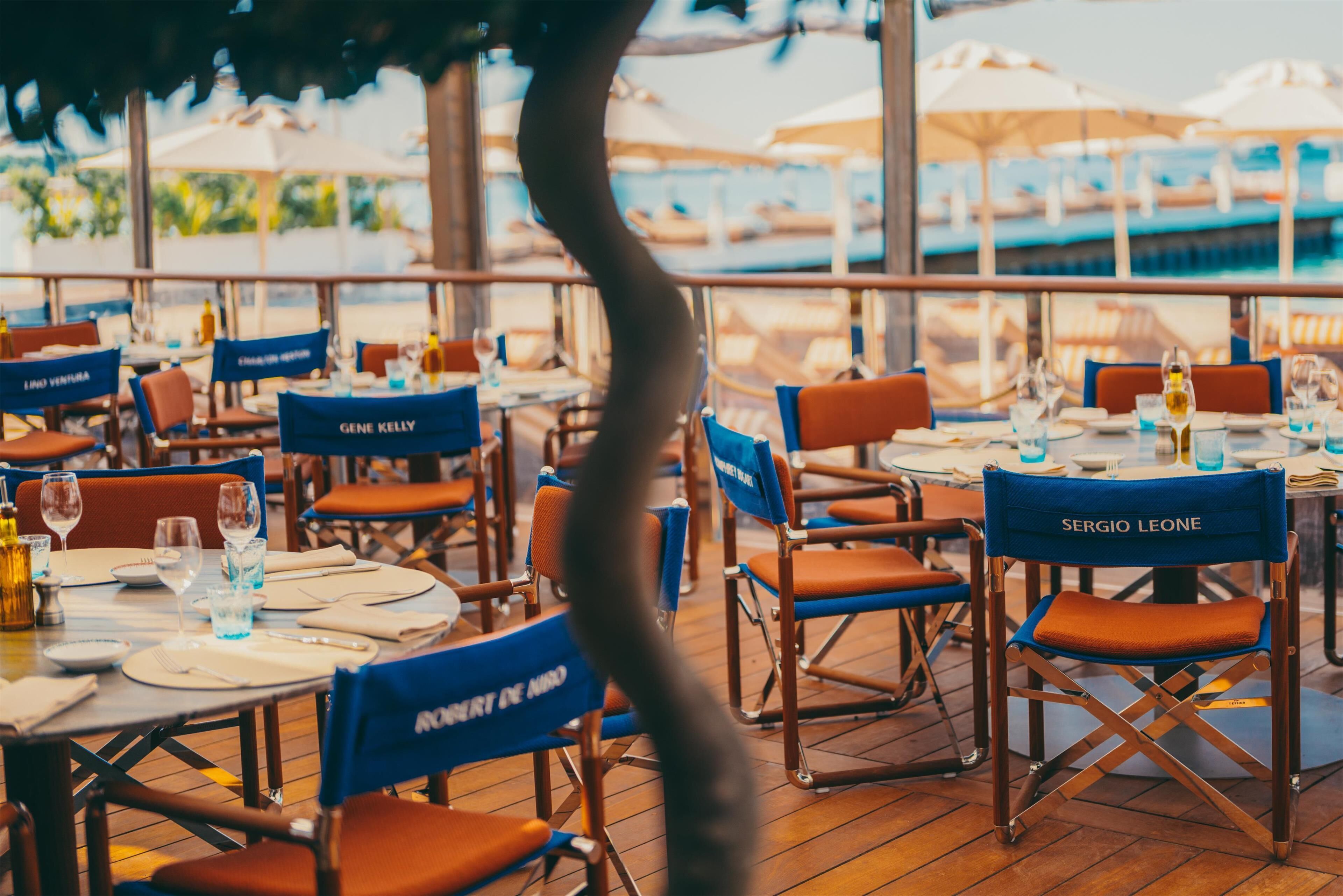 A restaurant with tables set for dining, chairs labeled with names, and a view of the sea through large windows.