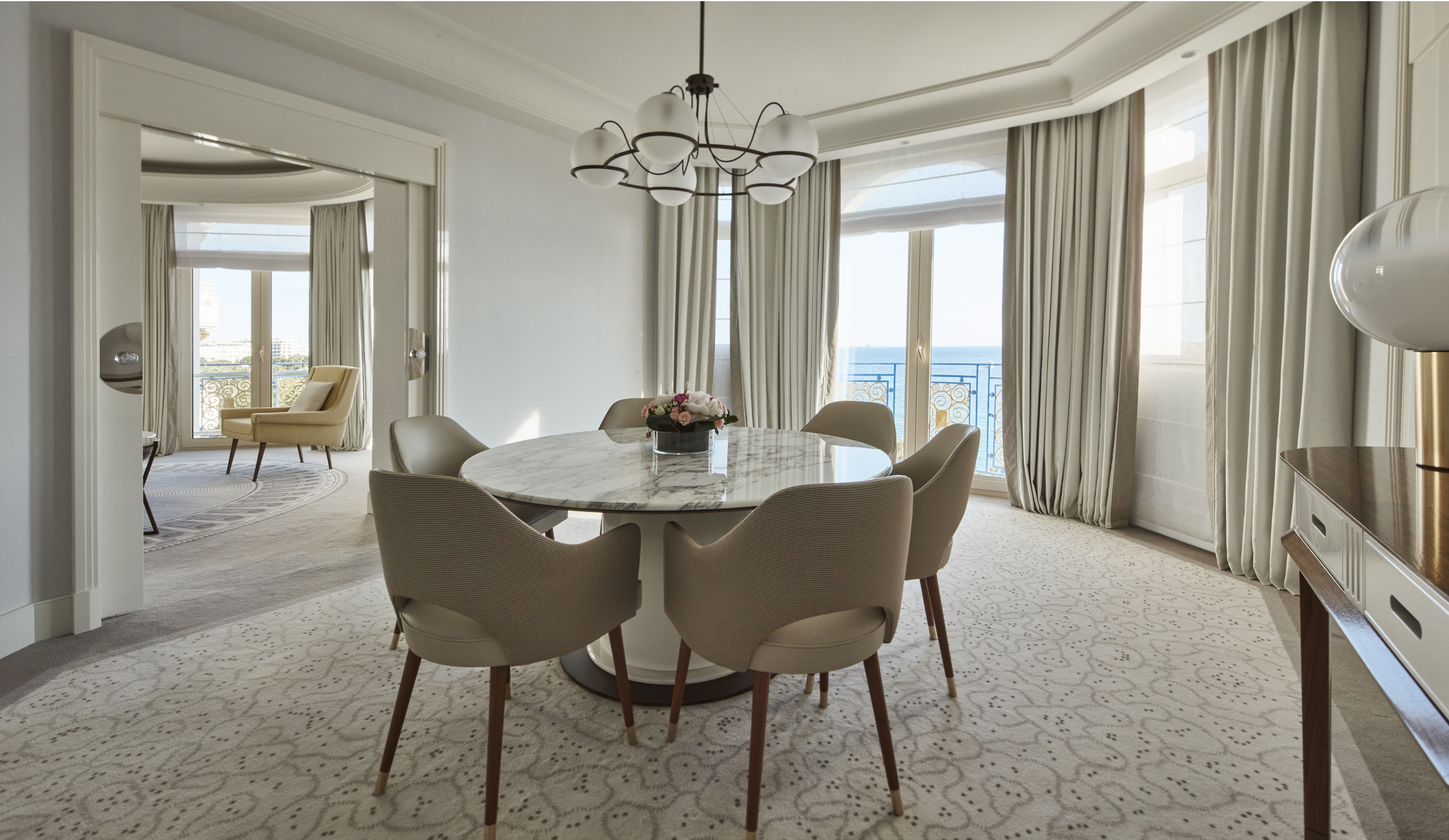 A hotel dining area with a round marble table, six wicker chairs, a chandelier, and a view of the sea through large windows.