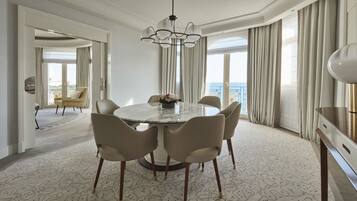 A hotel dining area with a round marble table, six wicker chairs, a chandelier, and a view of the sea through large windows.