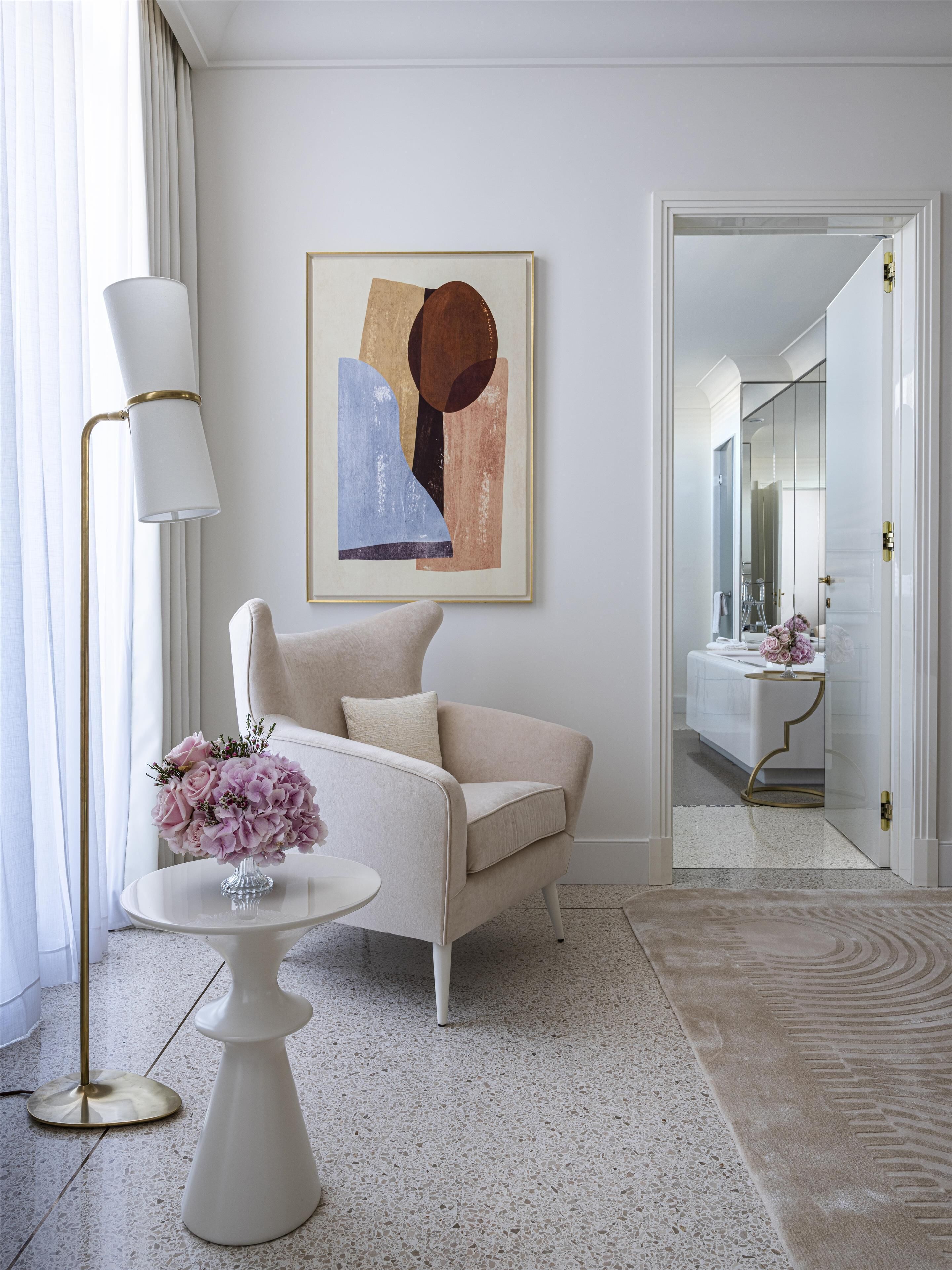 Una sala moderna con un sofá beige, una mesa redonda blanca con un jarrón de flores, una lámpara de pie y un cuadro abstracto enmarcado en la pared.