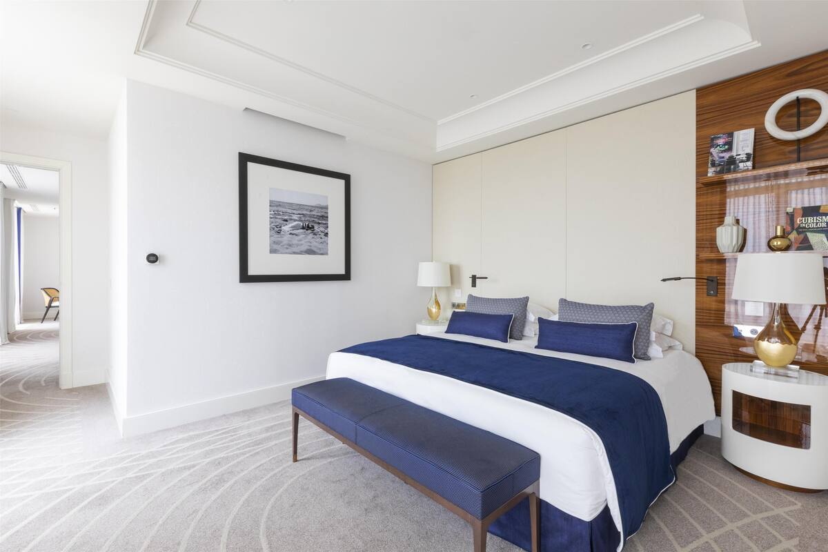 Une chambre d’hôtel moderne dotée d’un grand lit, d’une banquette, d’une table de chevet avec une lampe et d’une étagère murale agrémentée d’objets décoratifs.