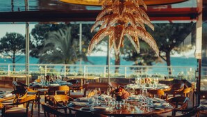 A restaurant with round tables set for dining, large windows offering a view of the sea, and a distinctive palm tree chandelier.