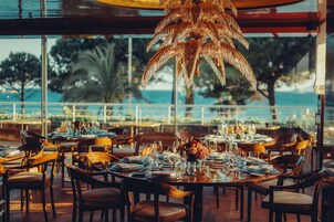 Een restaurant met ronde tafels gedekt voor diners, grote ramen met uitzicht op de zee, en een opvallende palmboomlichtkrans.
